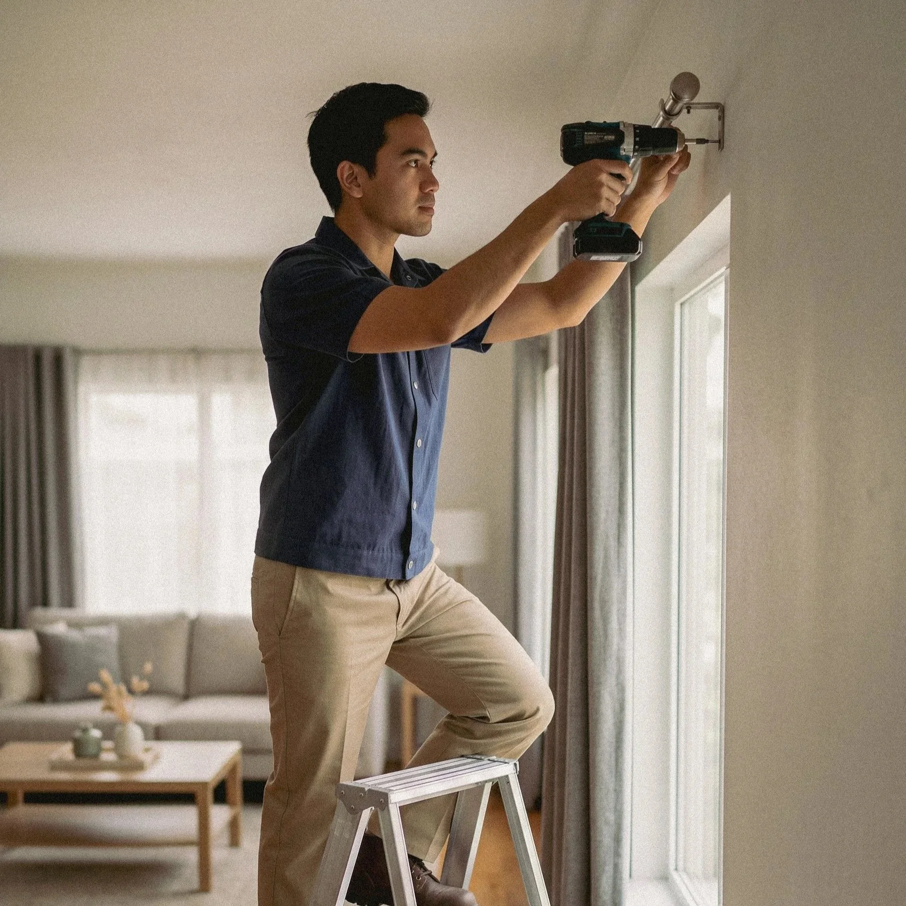 A man in a dark shirt and beige pants using a cordless drill to install a ceiling light fixture while standing on a small white step ladder in a well-lit living room with beige walls and curtains.