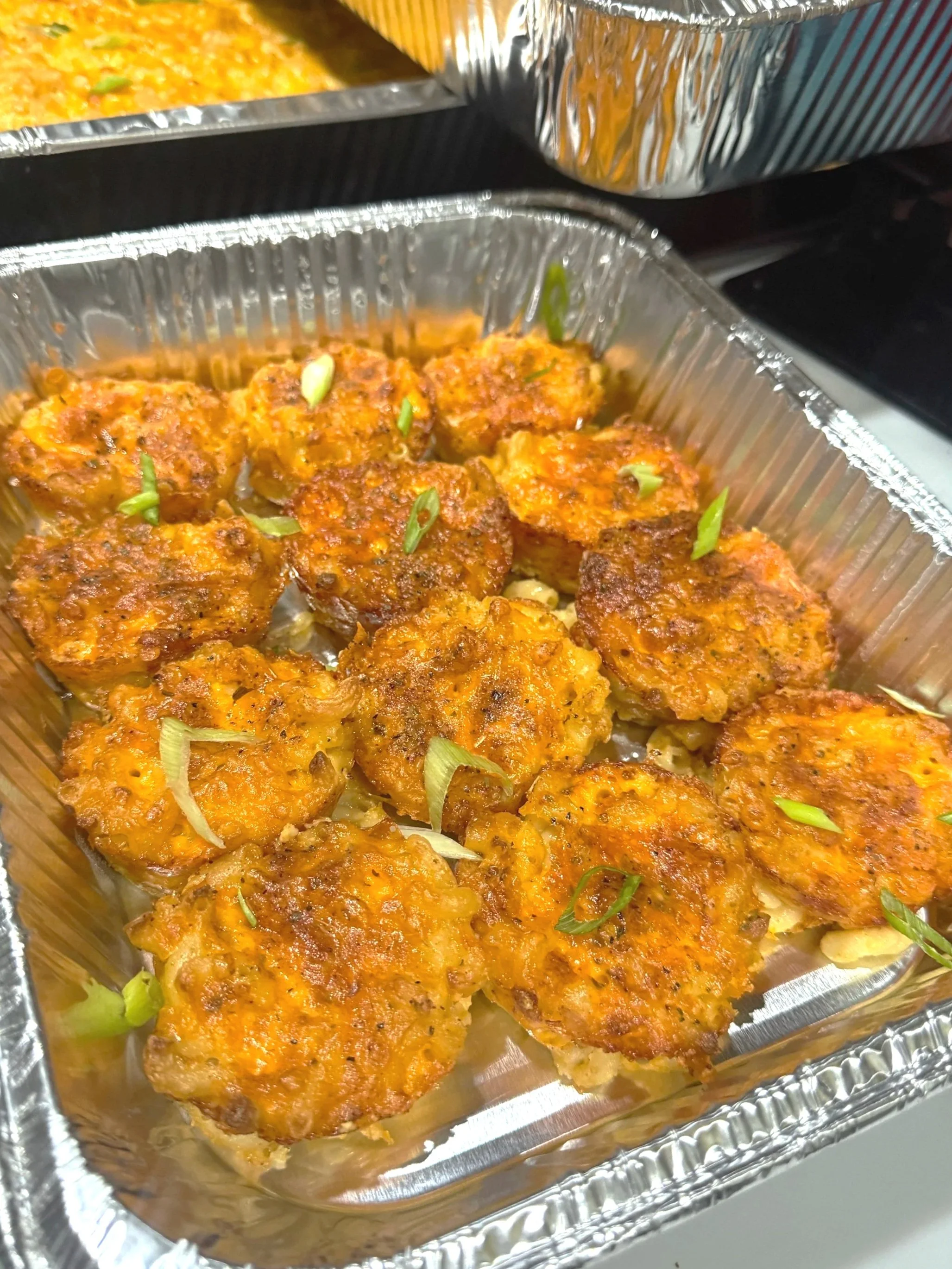 Aluminum tray filled with cooked meatloaf slices garnished with chopped green onions, in the foreground, with more trays of food in the background.