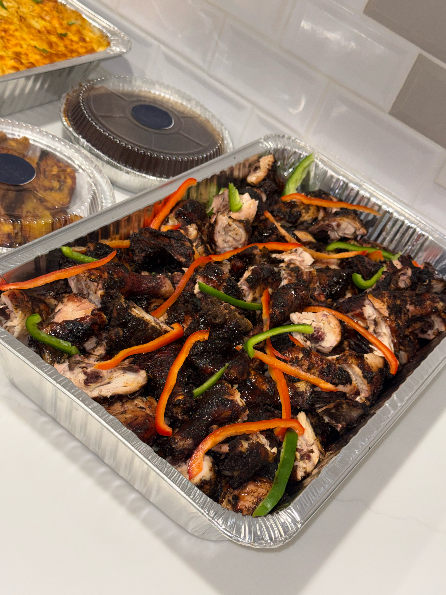 Tray of grilled chicken pieces garnished with sliced red and green peppers, with foil containers of food and sauce in the background.