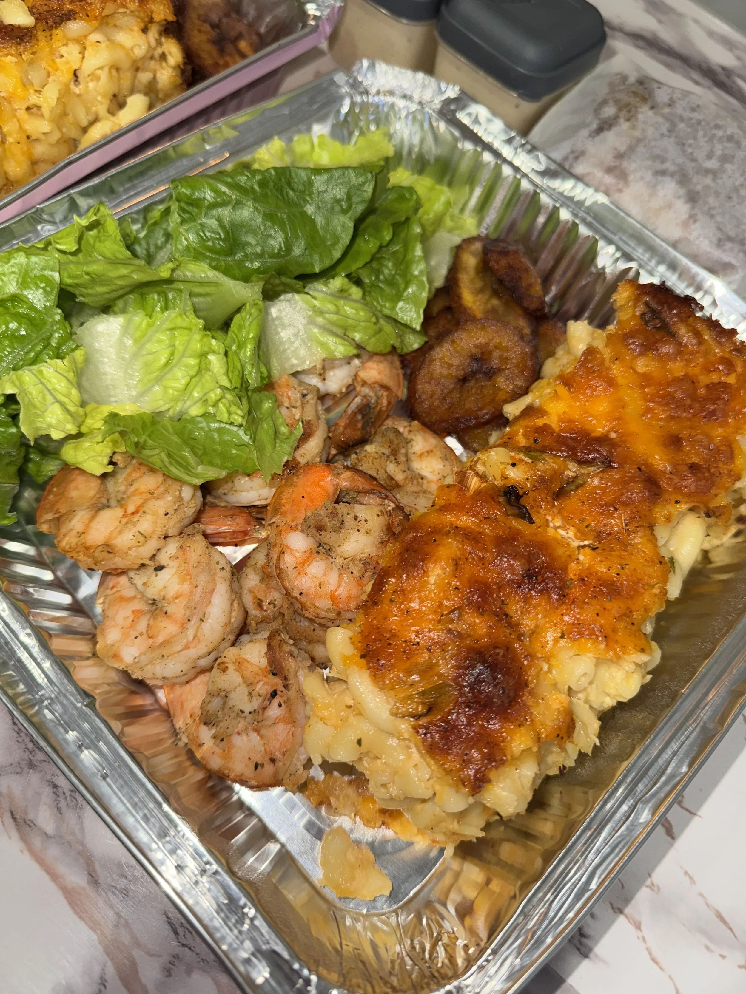 A foil container with a baked casserole topped with cheese and sauce, a side of cooked shrimp, and green lettuce leaves.