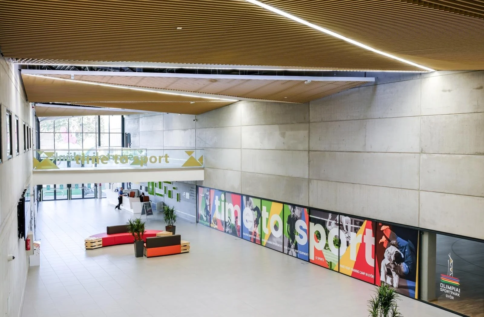 Interior of a modern sports complex with a sign that reads 'multisport' and a colorful promotional banner displaying images of athletes and text promoting sports activities. The space features beige tiled floors, concrete walls, and wooden ceiling pa