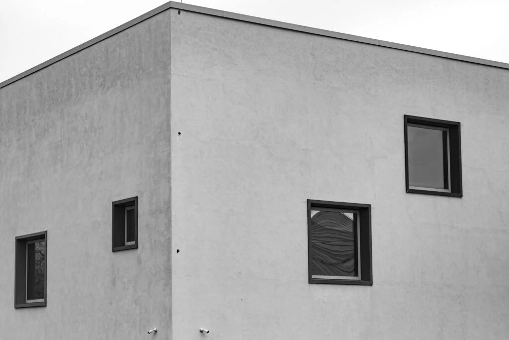 A black and white photo of a modern building with a flat roof, textured white exterior, and three rectangular windows with dark frames.