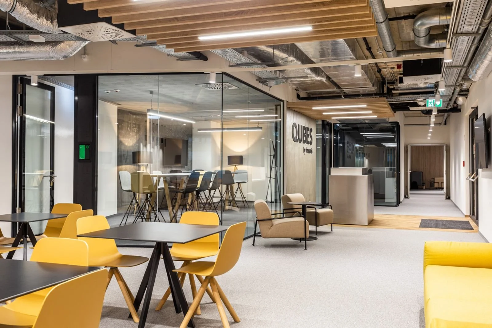 Modern office lounge with yellow chairs, black tables, and a glass enclosed meeting room labeled 'QUBES'. The space has wood and metal accents, with exposed ceiling pipes and a white and beige color scheme.
