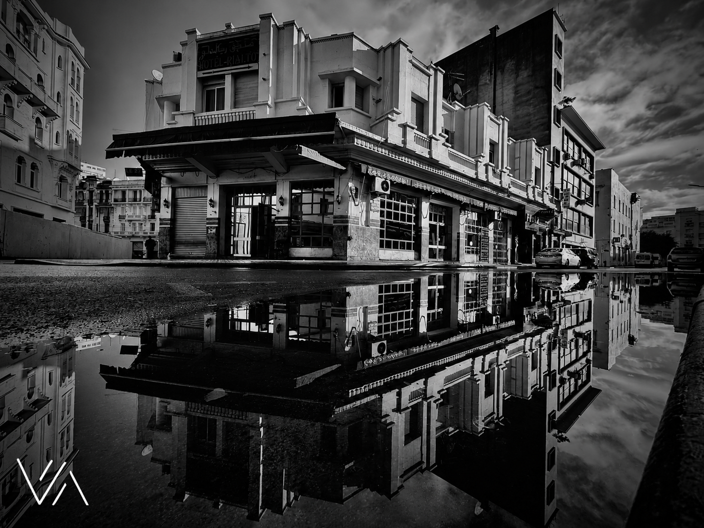 A black and white photograph of a city street reflecting a building and the sky in a large puddle, with cars parked along the curb and surrounding architecture.
