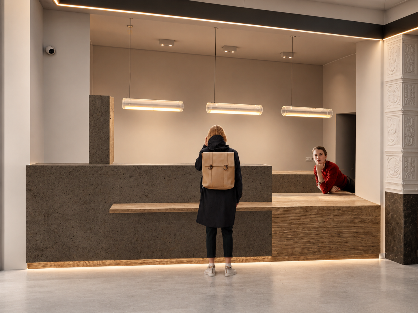 A woman with a beige backpack standing at a modern, minimalist reception desk in a contemporary building, with another woman in red leaning on the desk and looking towards the camera.
