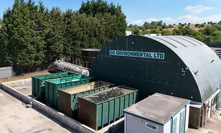 Large industrial building labeled 'Go Environmental Ltd' with several green waste containers outside on a concrete pad, surrounded by trees and hills in the background.