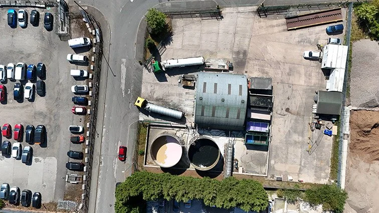 Aerial view of an industrial yard with large storage tanks, trucks, and parked cars, surrounded by a paved area and a curved road