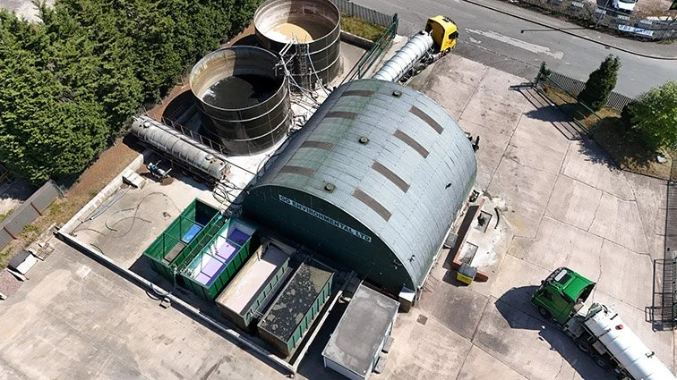 Aerial view of a waste management facility with multiple trash containers, a large curved-roof building, two large metal tanks, and several trucks parked on a concrete surface.