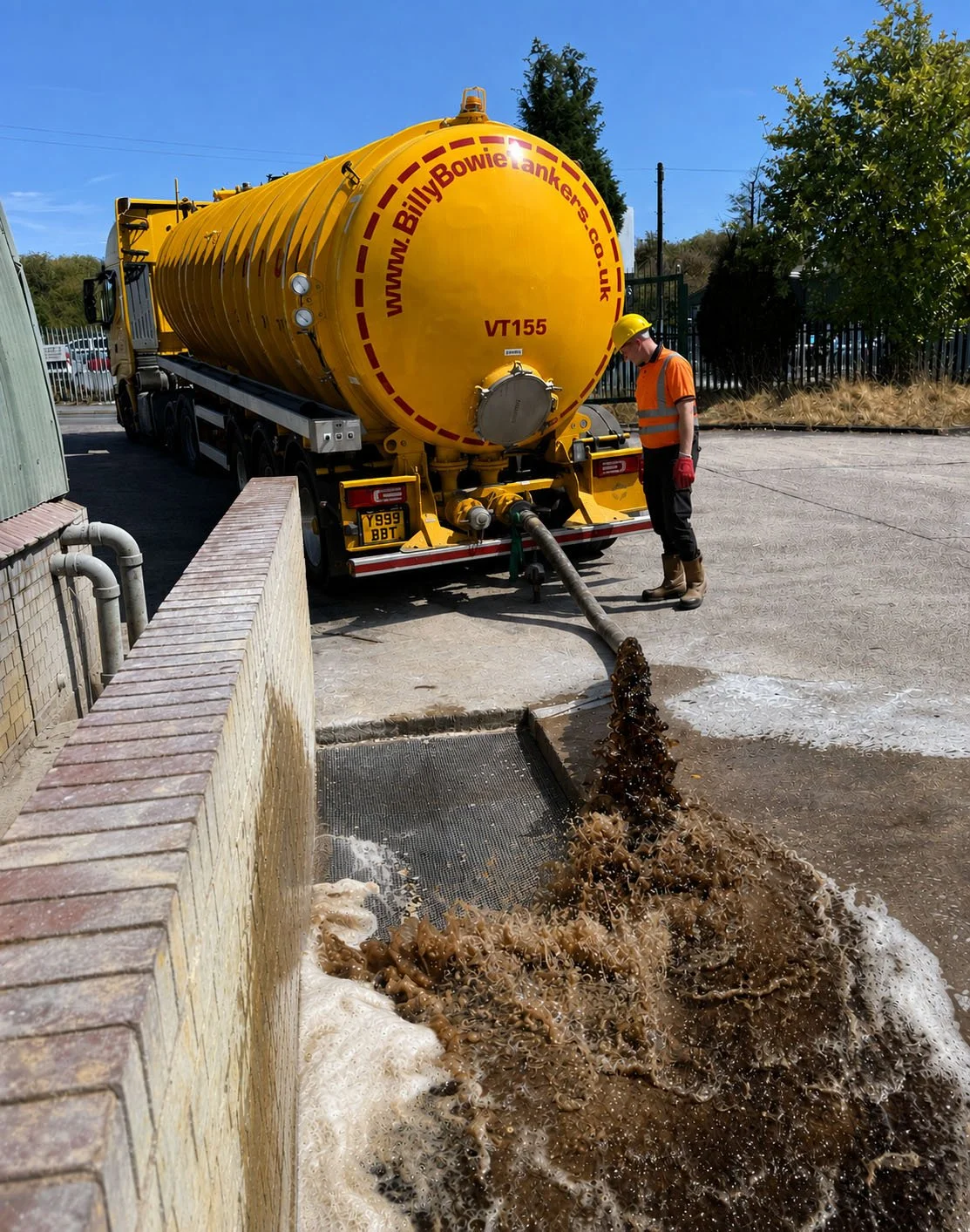 A worker in safety gear hoses a brown liquid substance from a yellow tanker truck into a drain.