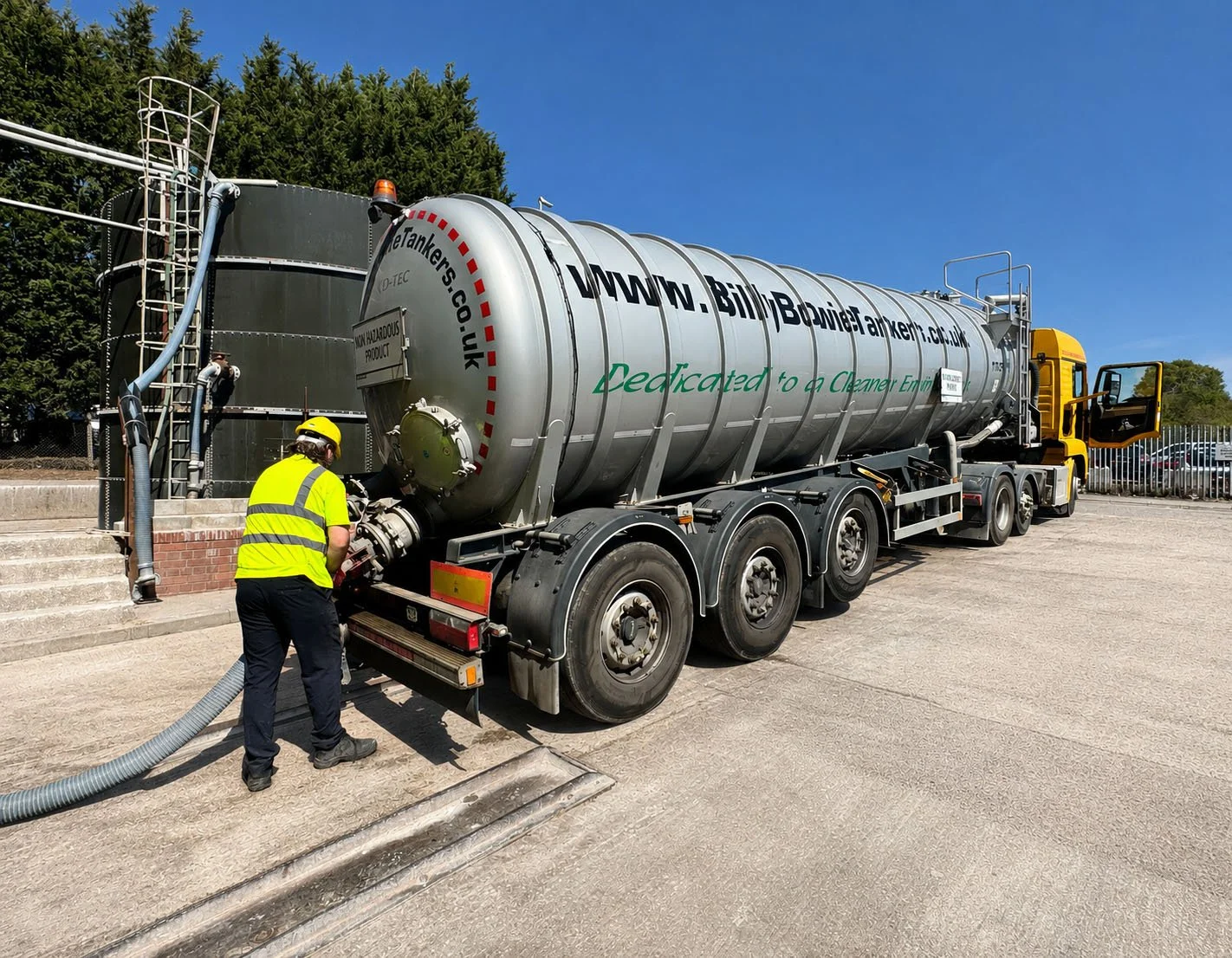 A worker in a yellow safety vest and helmet handling equipment near a large tanker truck labeled 'www.billitanker.co.uk,' with a ladder and pipes on the side, parked on a paved area outside, with trees and a blue sky in the background.