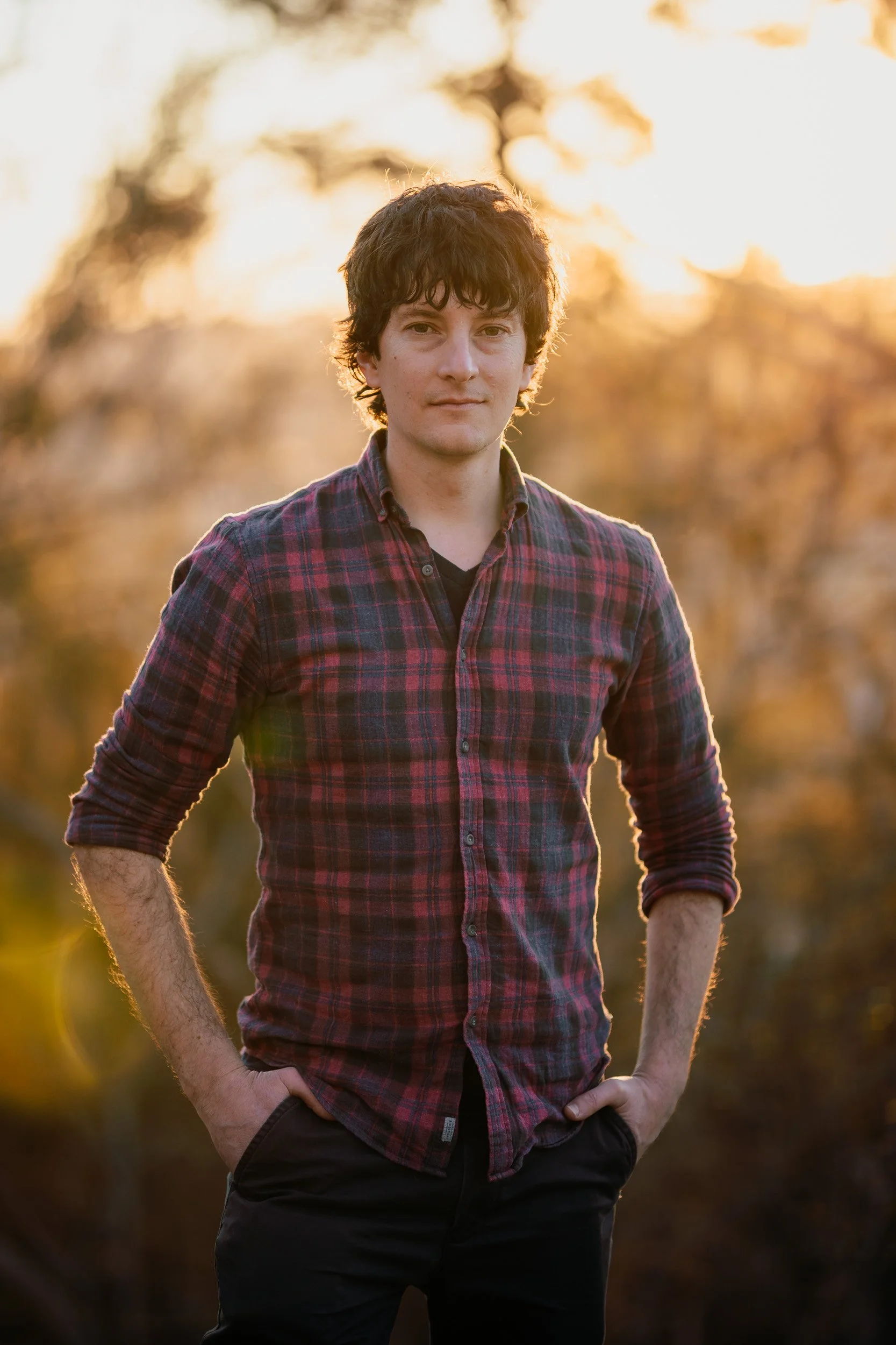 A young man with dark curly hair, wearing a red plaid shirt, standing outdoors during sunset with blurred trees in the background.