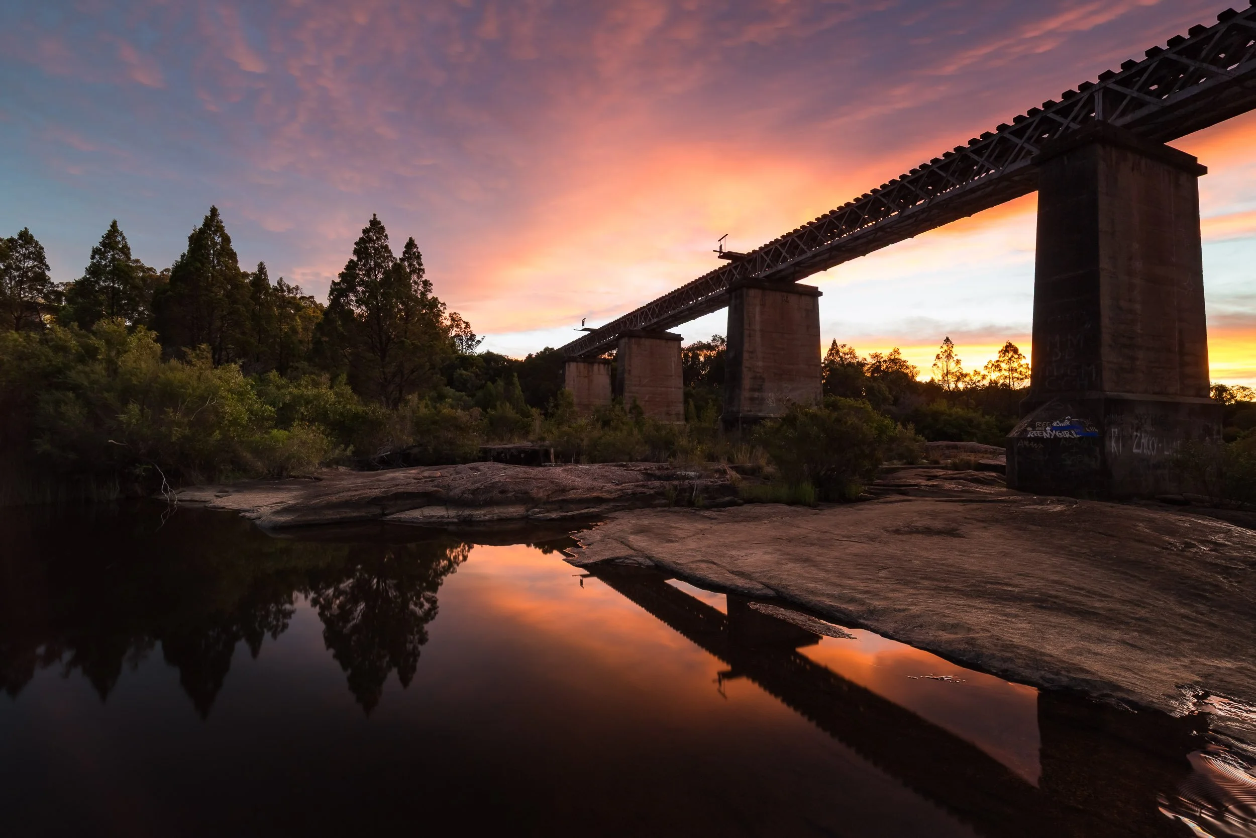 A sunset over Red Bridge at Quart Pot Creek in Stanthorpe with reflections of trees and a bridge, sky painted with pink and orange clouds.