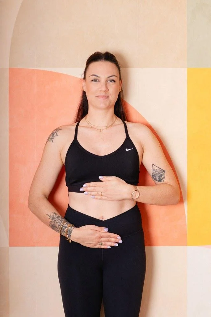 Femme debout, portant une tenue de sport noir, souriant, avec des tatouages sur le bras et l'épaule, devant un mur coloré.