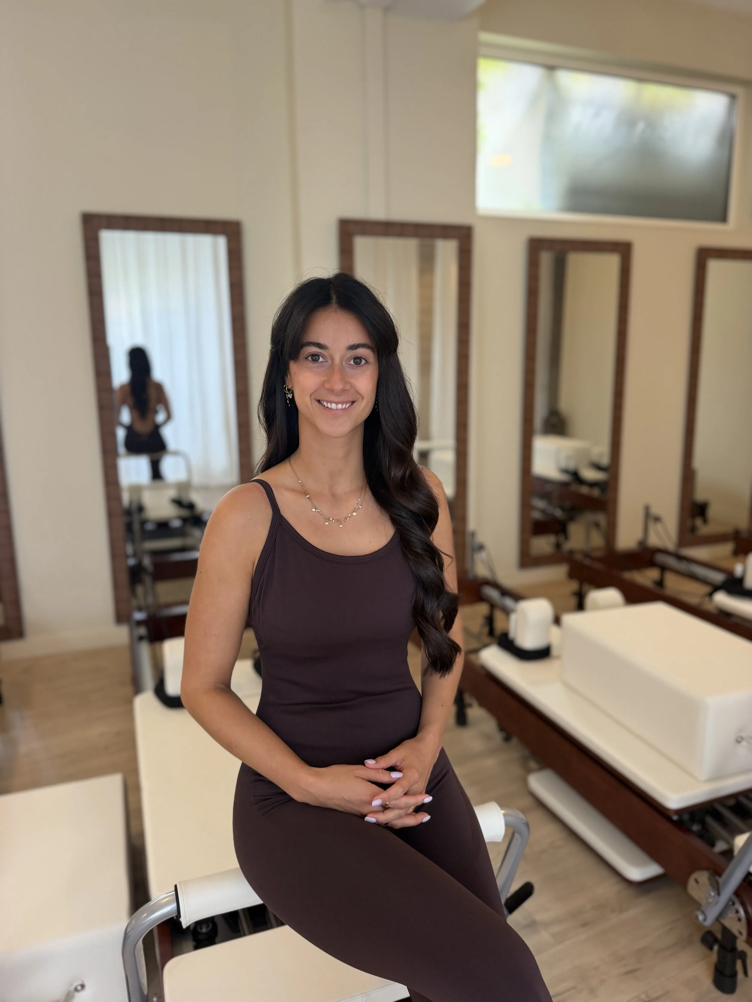 Une femme souriante avec des cheveux longs et noirs, portant une robe marron, posant dans un endroit lumineux avec plusieurs miroirs et appareils de beauté ou de maquillage en arrière-plan.
