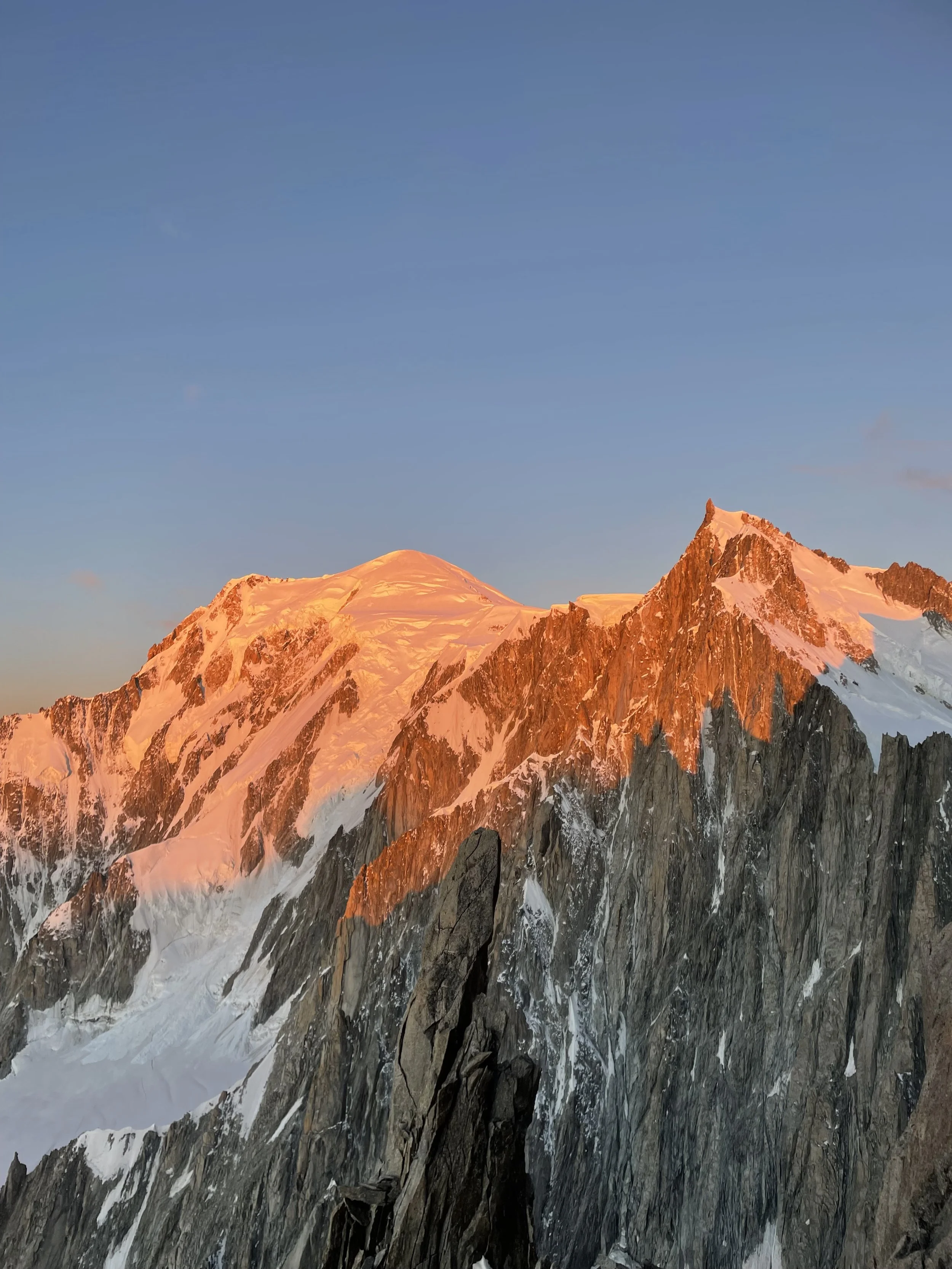 Montagne con cime innevate illuminate dal sole al tramonto, con cielo azzurro e poche nuvole.