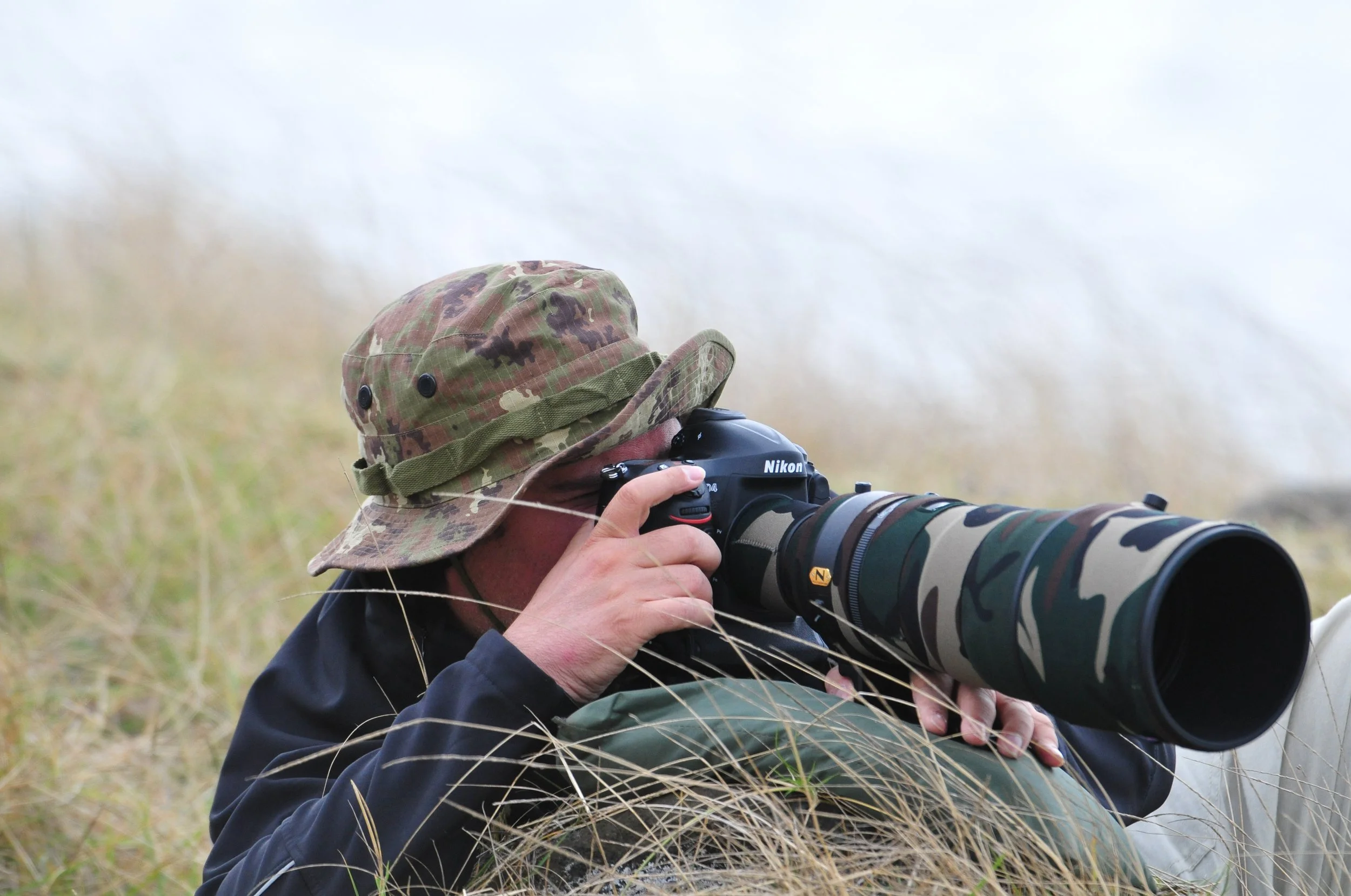 Io in appostamento con cappello mimetico mentre utilizzo un teleobiettivo Nikon per fotografare in un campo aperto con erba alta, durante una giornata nuvolosa.