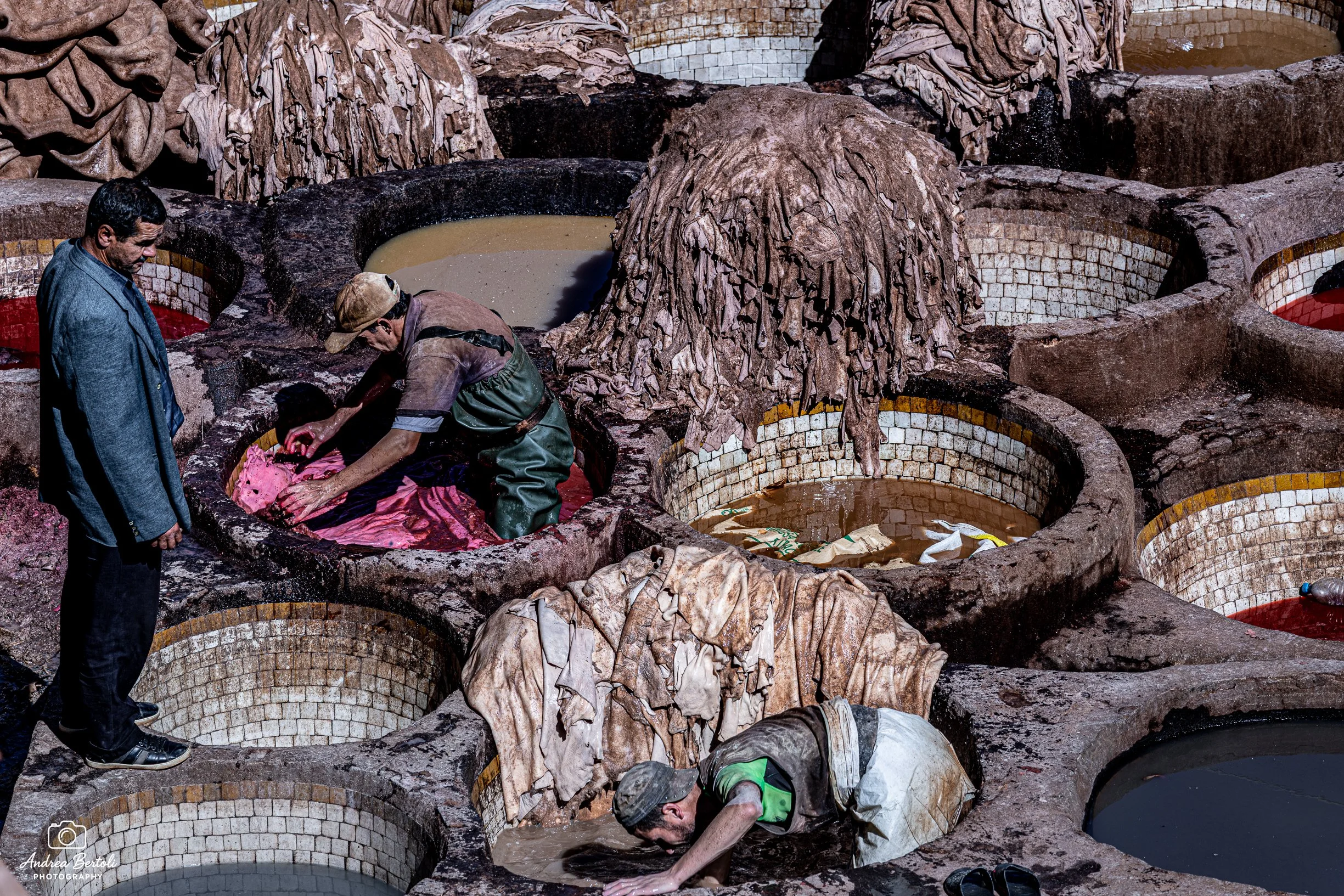 Personaggi intenti a lavorare con materiali organici nei grandi contenitori di un sito di conciatura delle pelli nella medina di Fès, con tubi e vasche di varie dimensioni e colori, ambientato all'aperto.