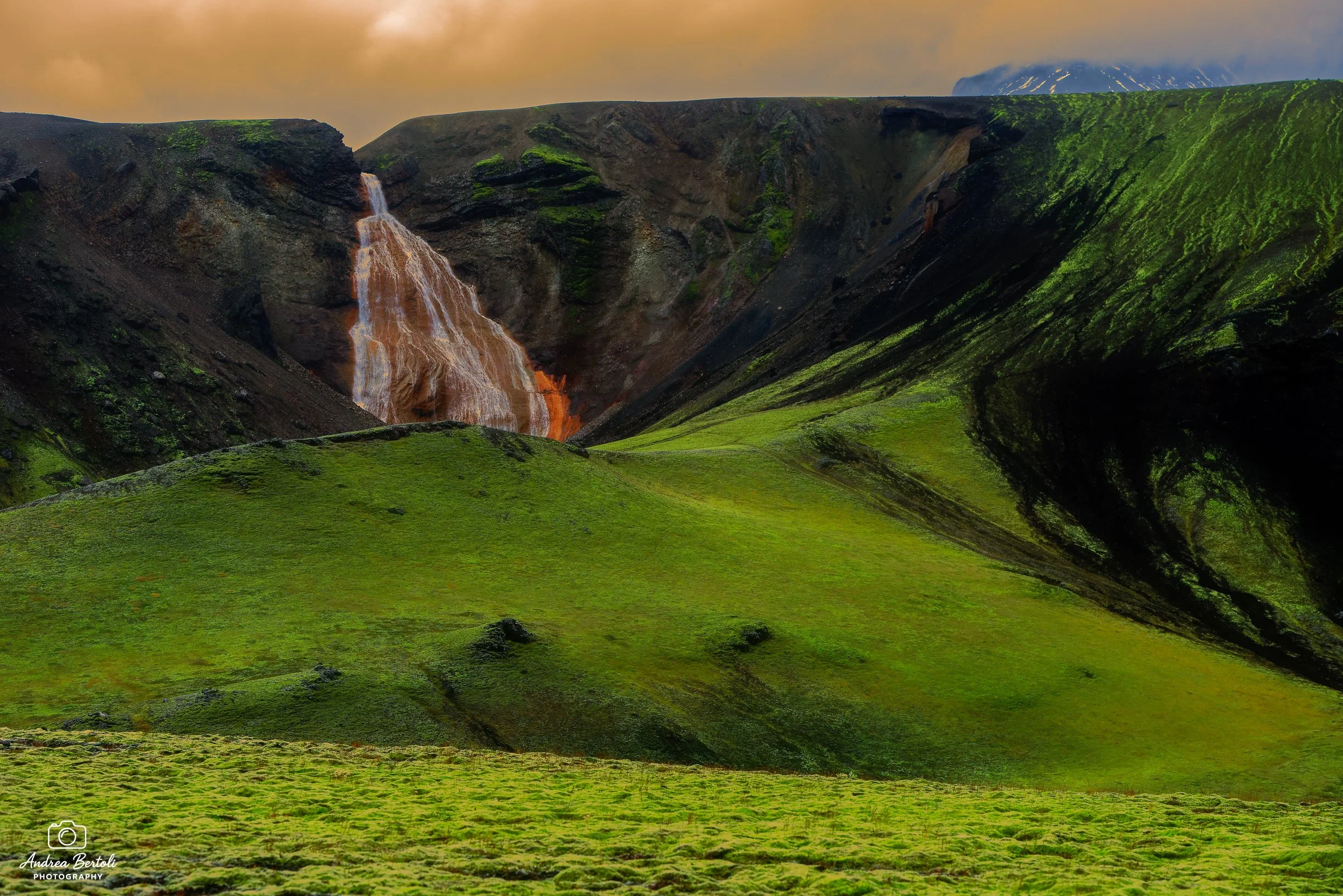 Paesaggio islandese con cascata, verdi colline e una montagna in lontananza sotto un cielo nuvoloso