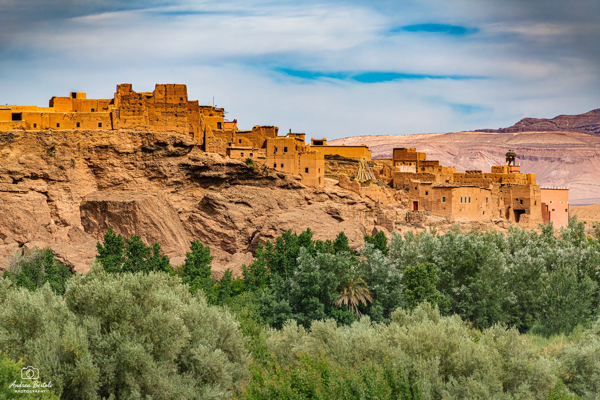 Antica città in terra arida con edifici in mattoni di adobe e mura in rovina lungo la Strada delle Mille Kasbah, circondata da vegetazione verde, con montagne sullo sfondo e cielo nuvoloso.