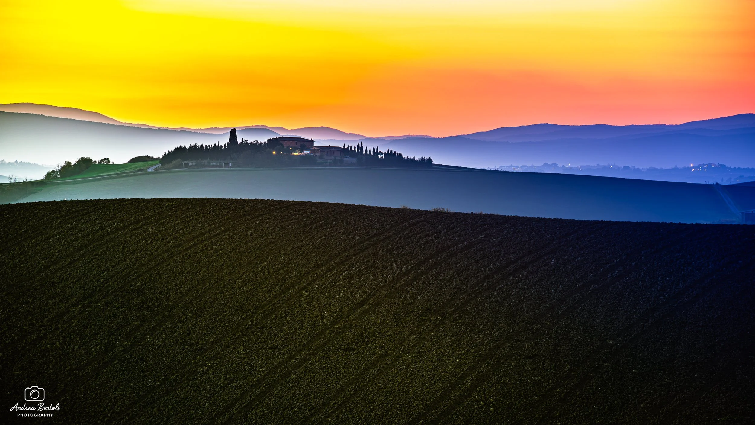 Paesaggio collinare senese al tramonto con cielo colorato di giallo, arancione e rosa, colline verdi e una casa isolata in cima a una collina