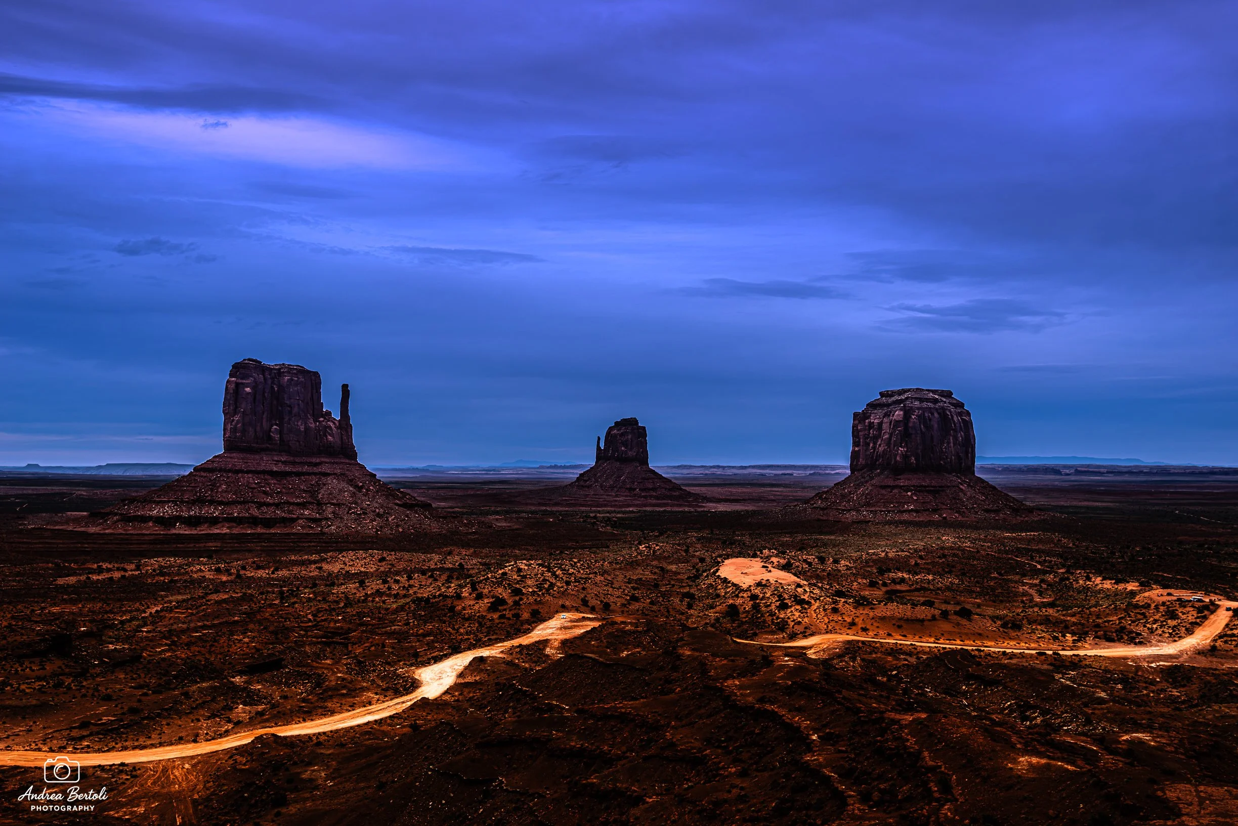 Vista della Monument Valley con tre grandi formation rocciose sotto un cielo nuvoloso, tramonto o alba con luci calde sul terreno.