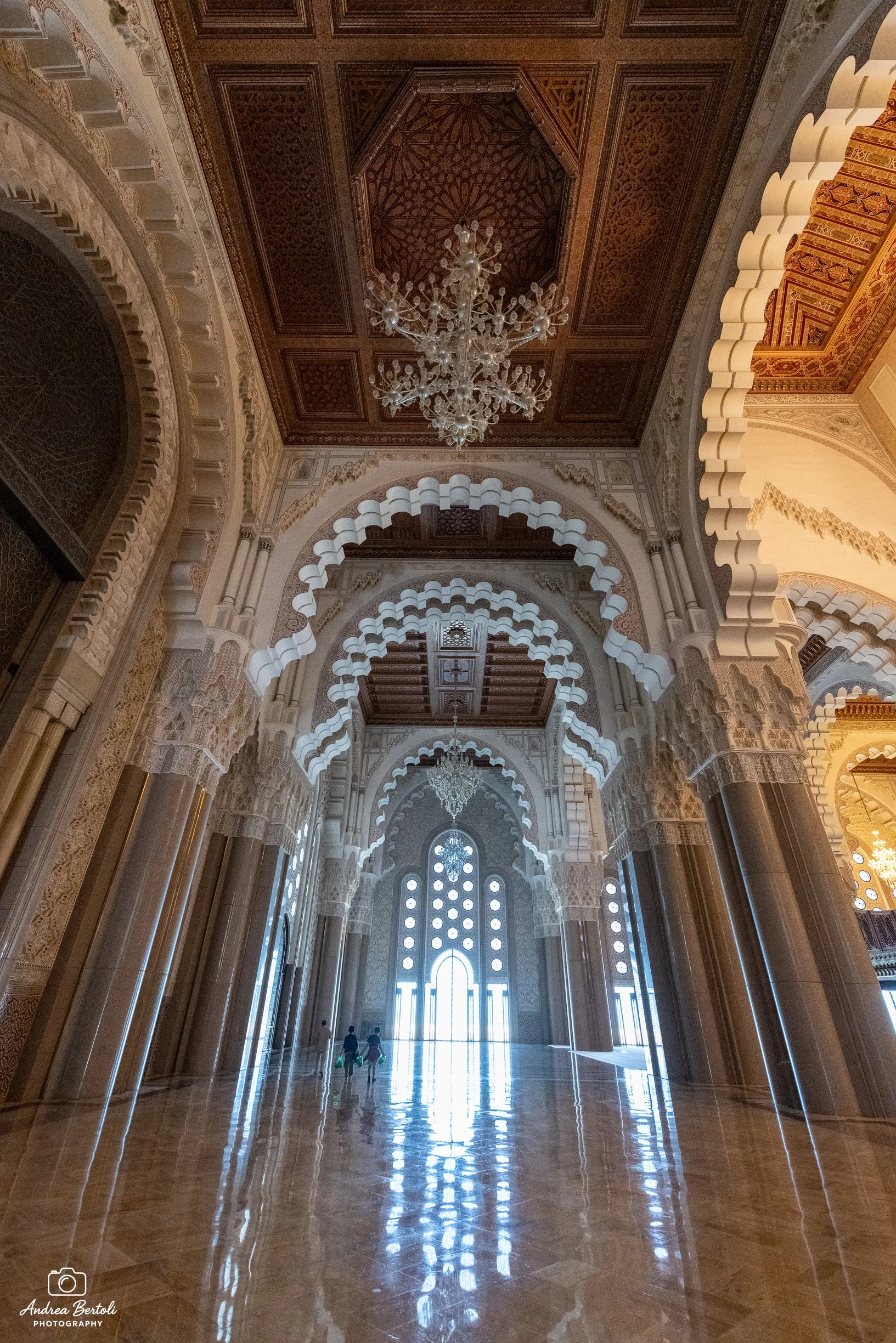 Interno della moschea Hassan II a Casablanca con Archi e colonne in marmo, grande lampadario di cristallo e finestre con motivi geometrici. Persone in lontananza che camminano lungo il corridoio.