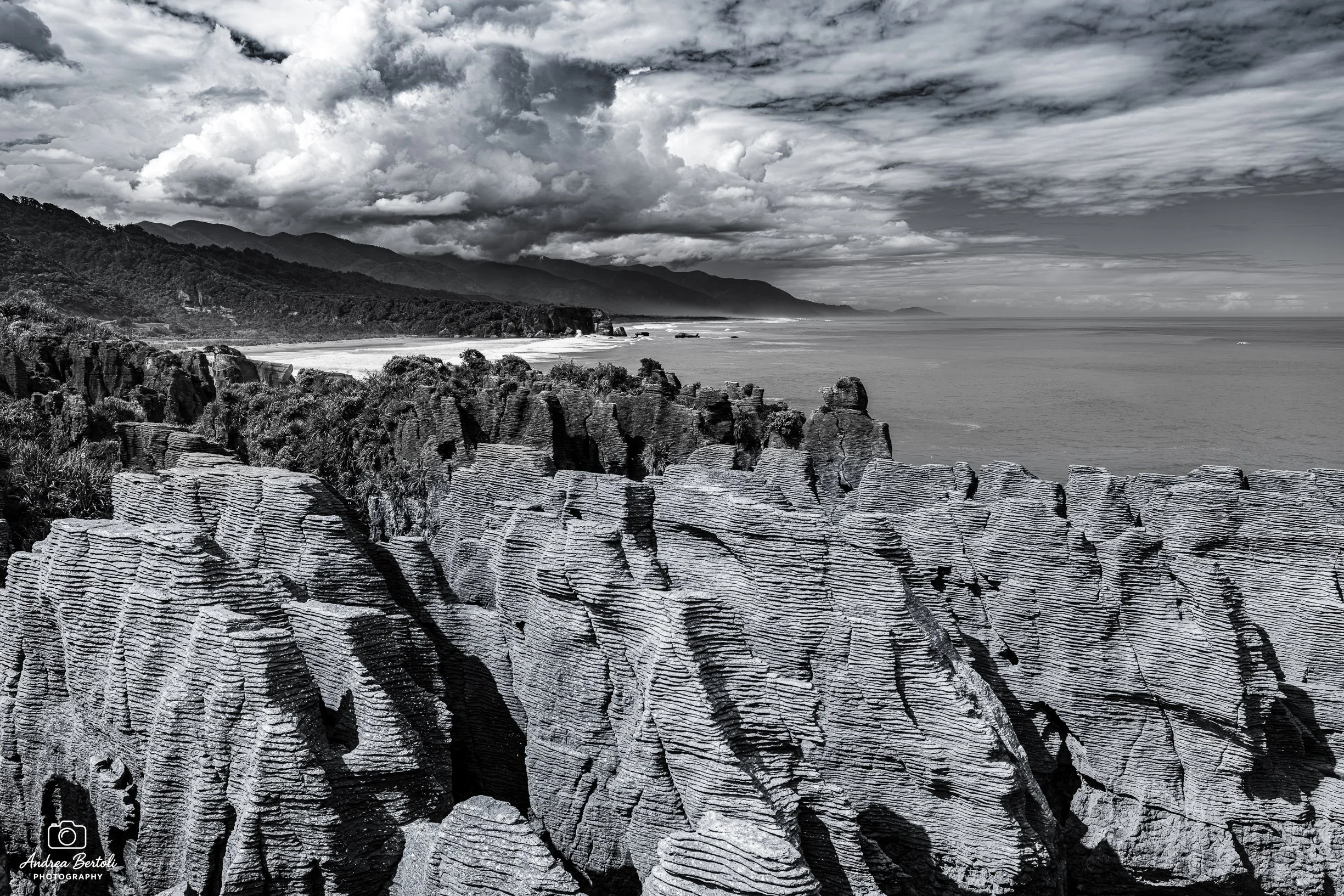 Paesaggio costiero con le Pancake Rocks in primo piano, mare e montagne sullo sfondo, nuvole dense nel cielo in bianco e nero.
