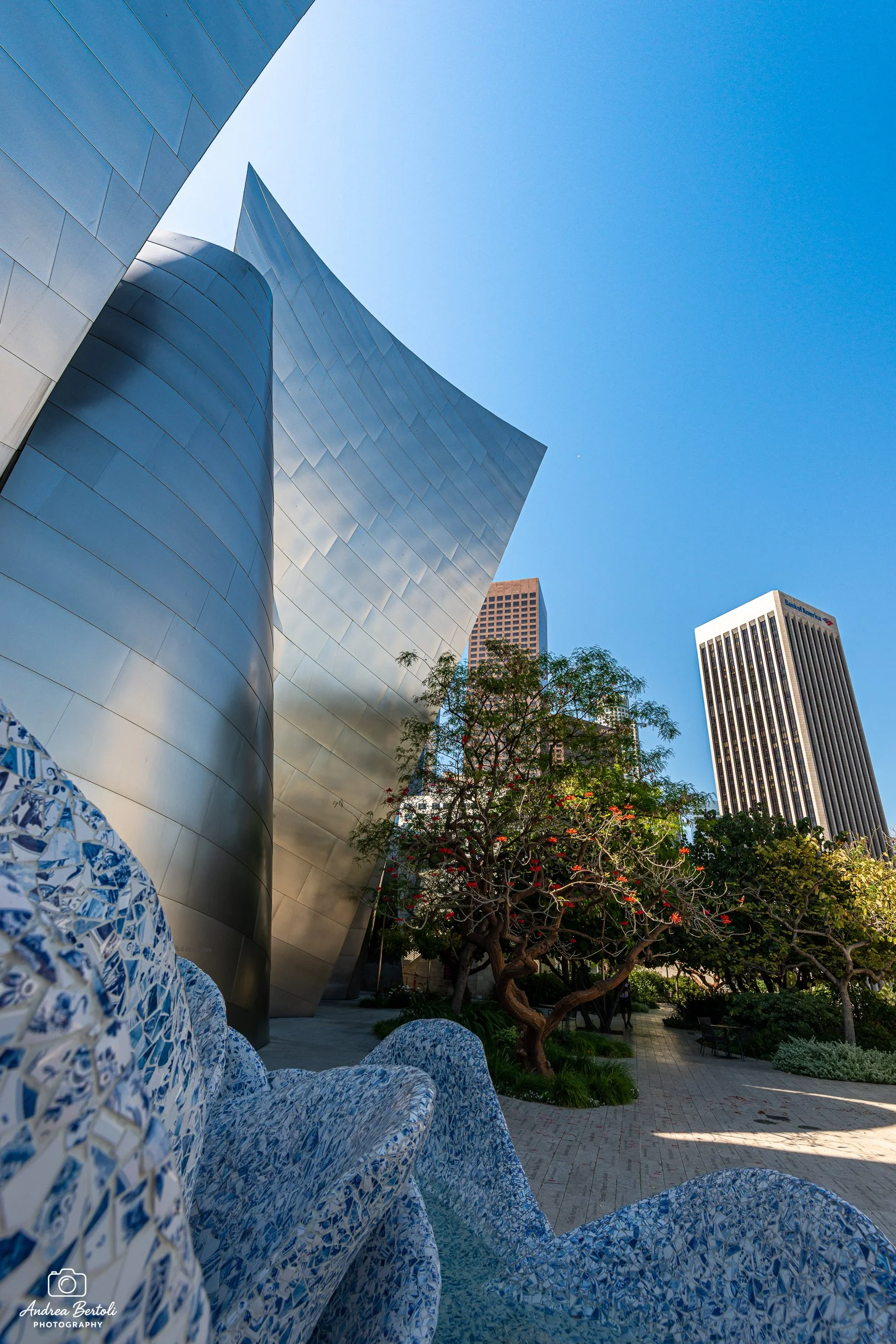 Vista della piazzetta interna del Walt Disney Concert Hall, alberi con foltage verde e fiori rossi, panchine e una passeggiata pedonale, cielo azzurro limpido.