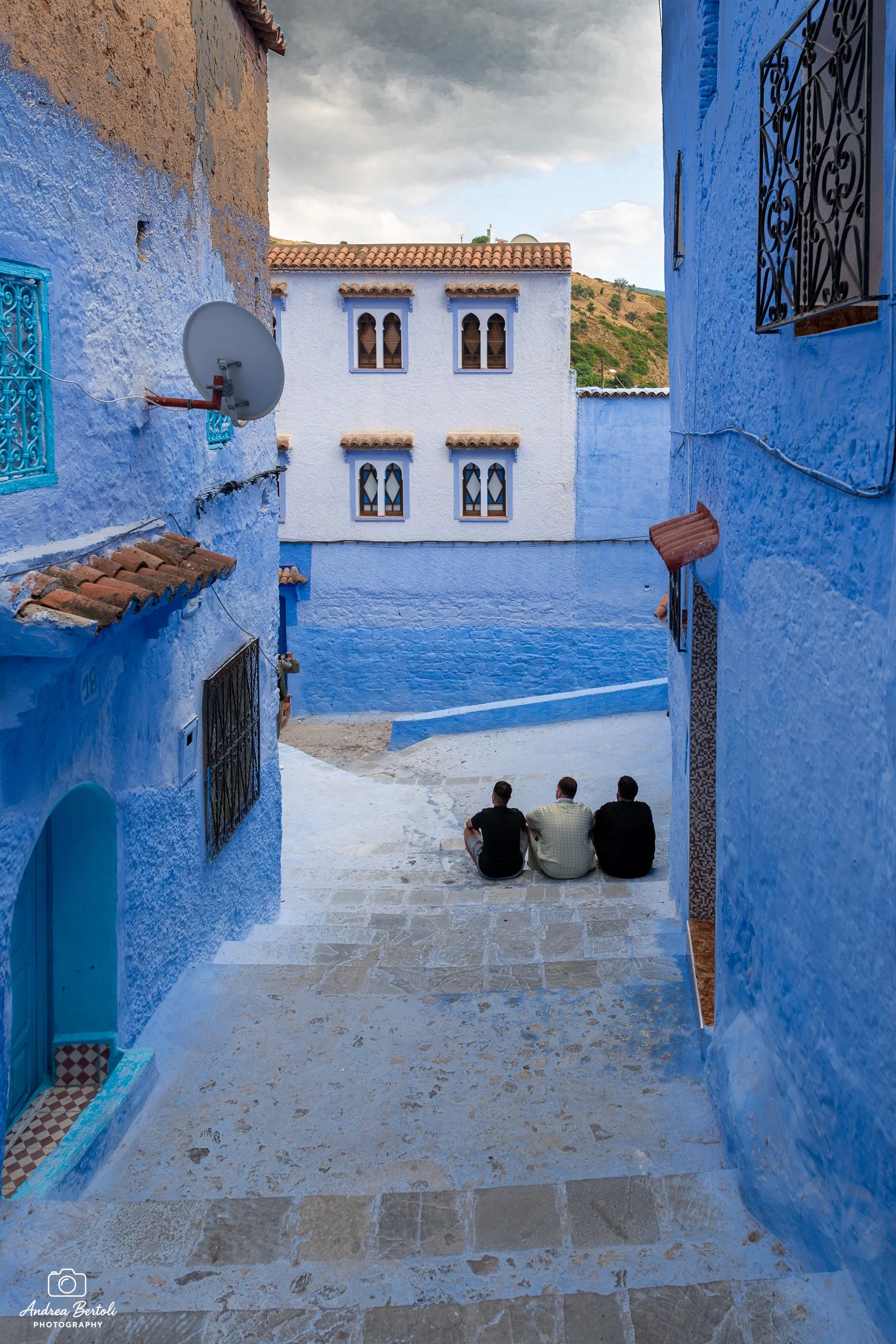 Tre persone sedute su un'area pavimentata in una piazzetta di Chefchaouen, circondata da edifici con muri blu e finestre con grate nere. In lontananza si vedono colline verdi con alcune case sparse.