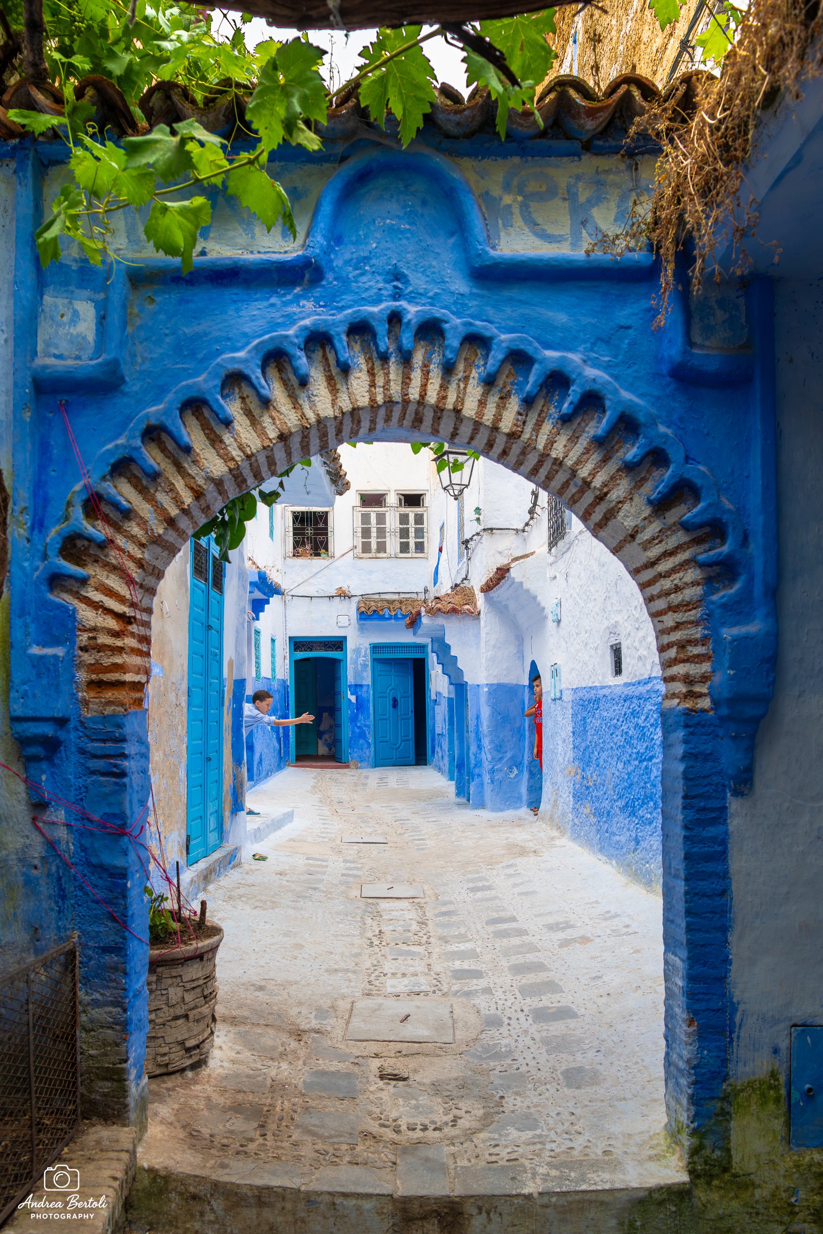 Una strada di Chefchaouen con case bianche e porte blu, vista attraverso un arco decorato con pittura azzurra e mattoni. Due bambini giocano sulla strada. Sul lato sinistro, ci sono piante in vaso.