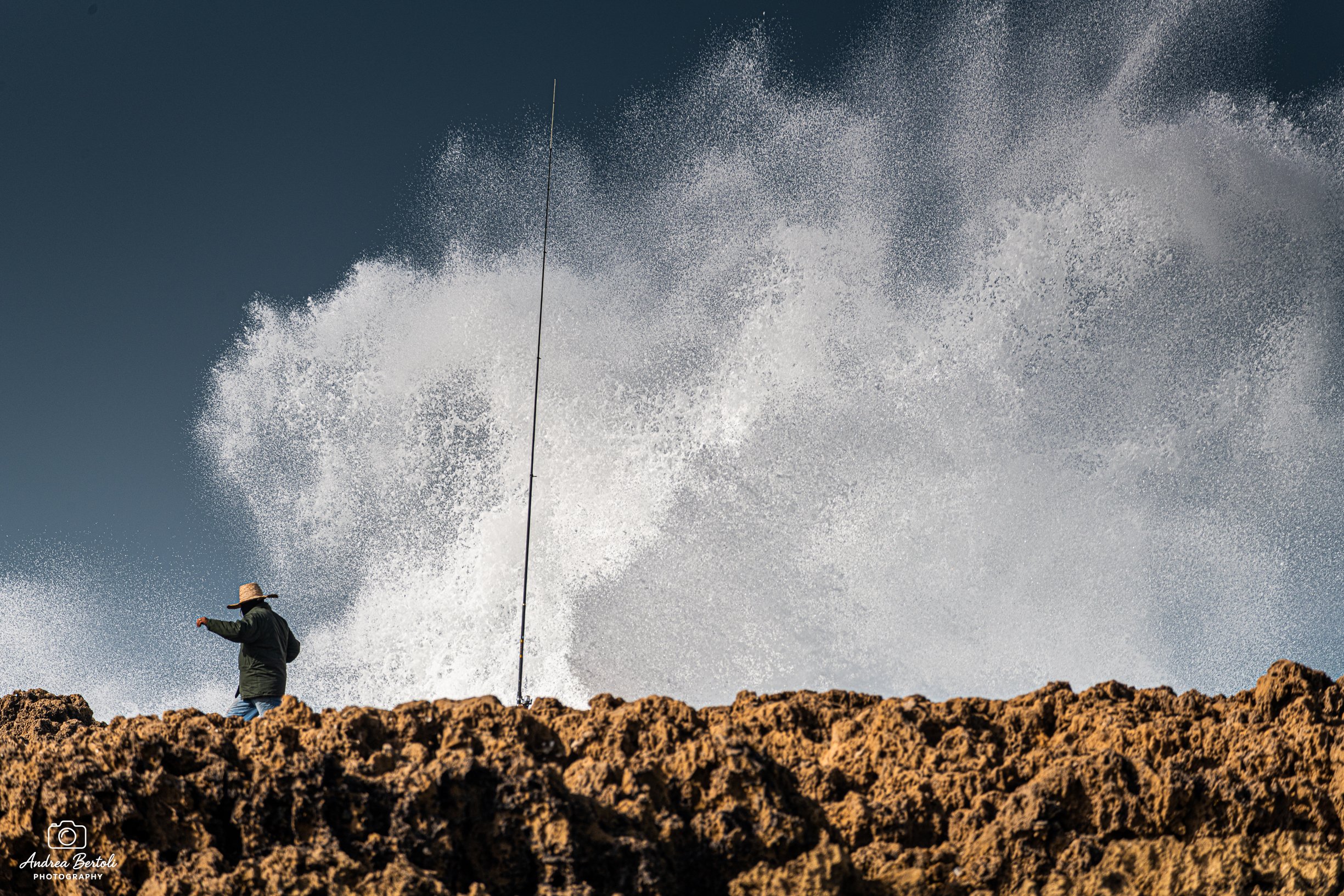 Un pescatore abbandona la sua canna da pesca per sfuggire all'arrivo di una grossa onda che sta per infrangersi sulla scogliera.