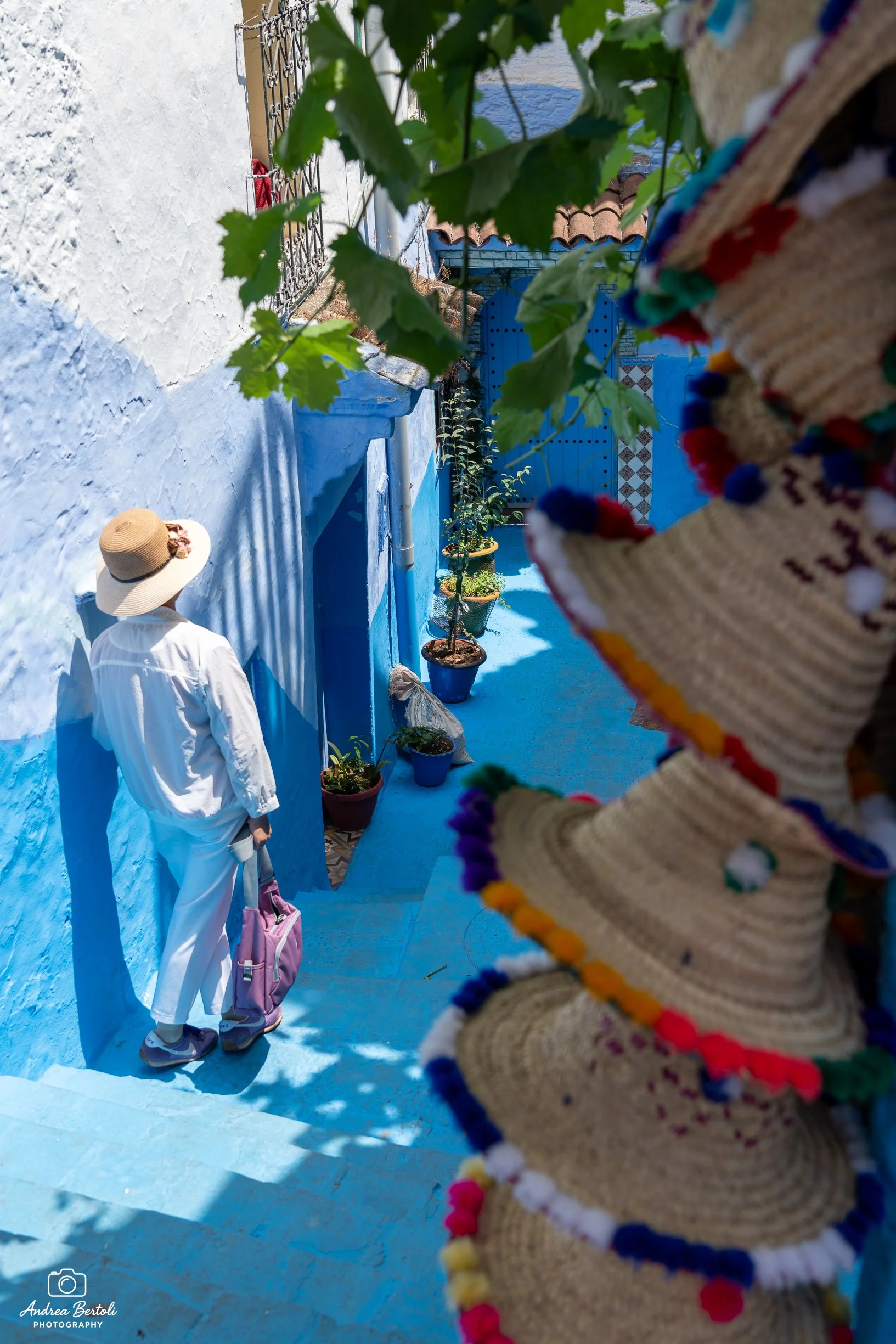 Una donna con cappello di paglia, vestita di bianco, con uno zaino rosa, cammina lungo una strada di Chefchaouen. La scena è animata da piante verdi e un muretto con bandiere colorate, con un arco in mattoni e una porta blu sullo sfondo.