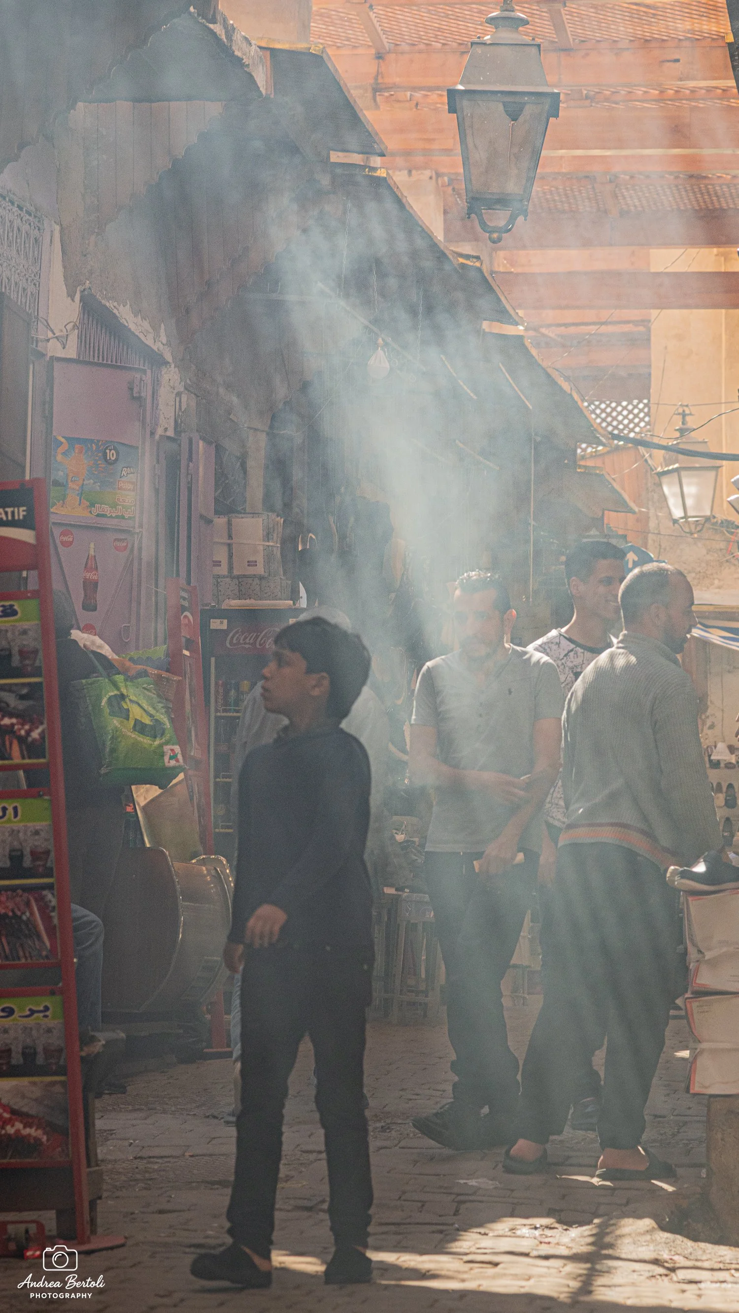 Il filmato mostra una scena di strada di giorno con alcune persone che camminano tra le bancarelle o i negozi di un mercato. La scena è leggermente avvolta dal fumo, e ci sono luci da lanterne o lampioni che illuminano la scena.