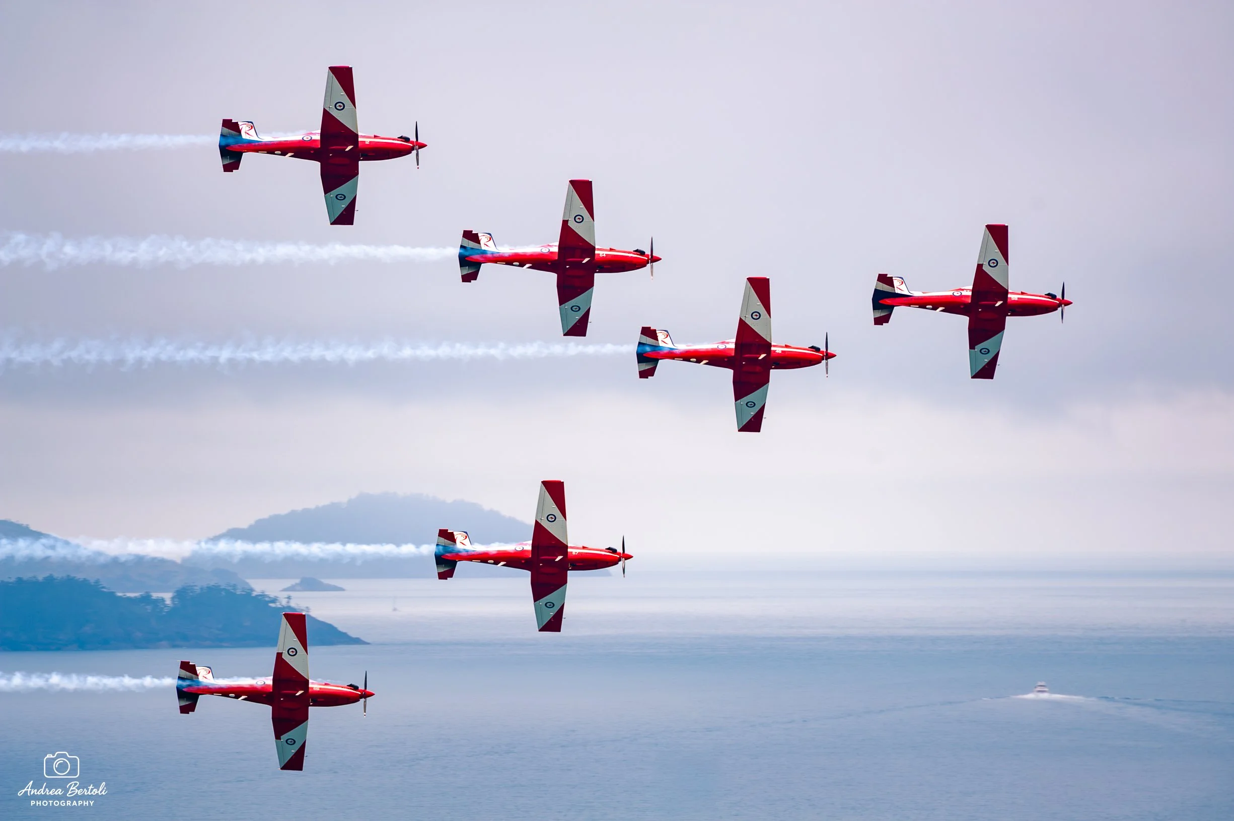 Sei aerei da acrobazia rossi in volo in formazione sopra il mare, con montagne e isole sullo sfondo, lasciando scie di condensa nel cielo.