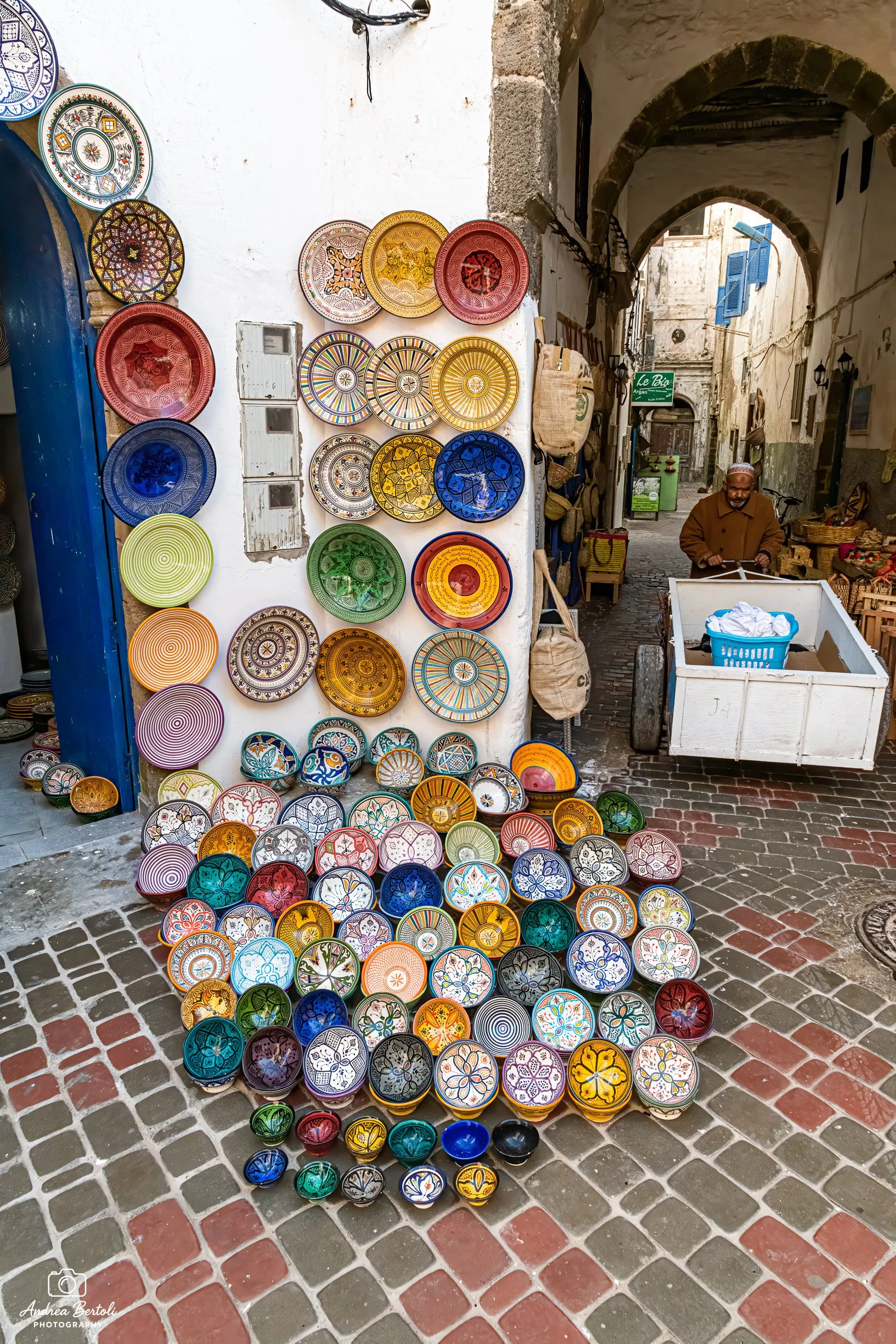 Vendita di piatti colorati in ceramica decorativa in un vicolo stretto con arcate, con un venditore anziano in piedi vicino a un carrello.