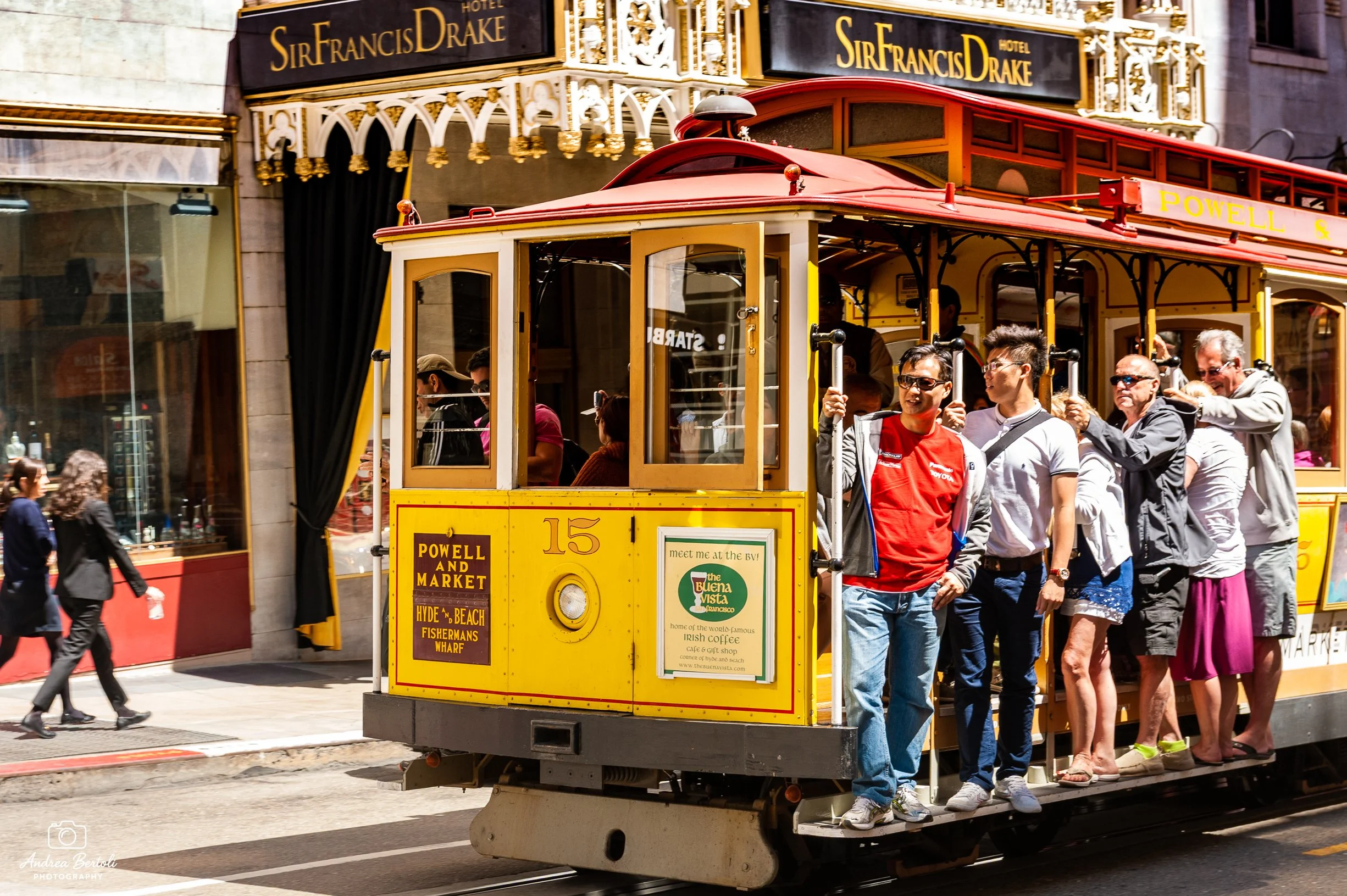 Un tram giallo e rosso con persone a bordo attraversa una strada in una zona affollata della città. Sul tram ci sono uomini e donne che siedono e si tengono per mano, alcuni sorridono. Sul retro si vede un grande cartellone con il nome dell'hotel Sir