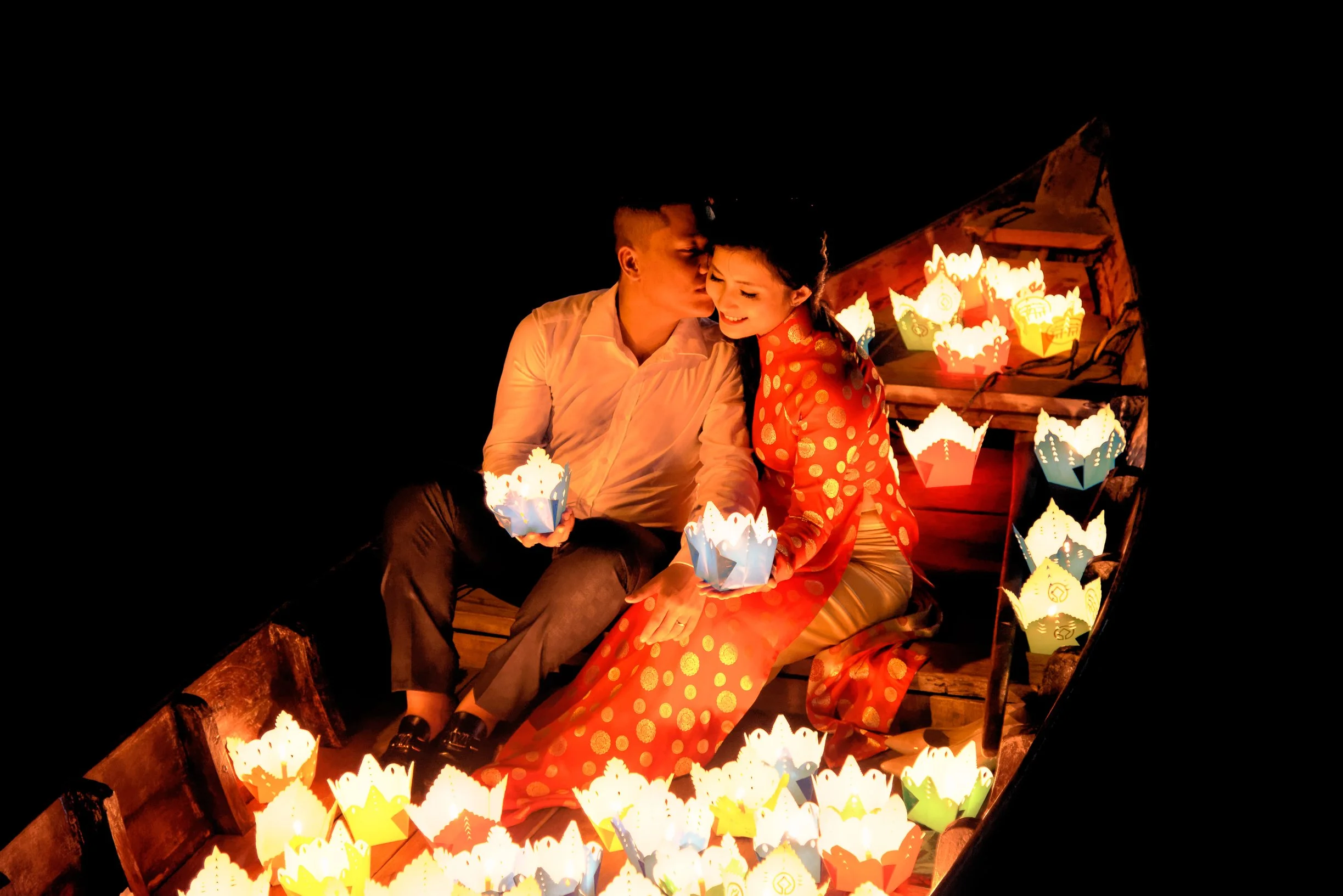 Una coppia di persone giovani sorride ed è abbracciata, davanti a vari lumini di carta colorati mentre è notte.