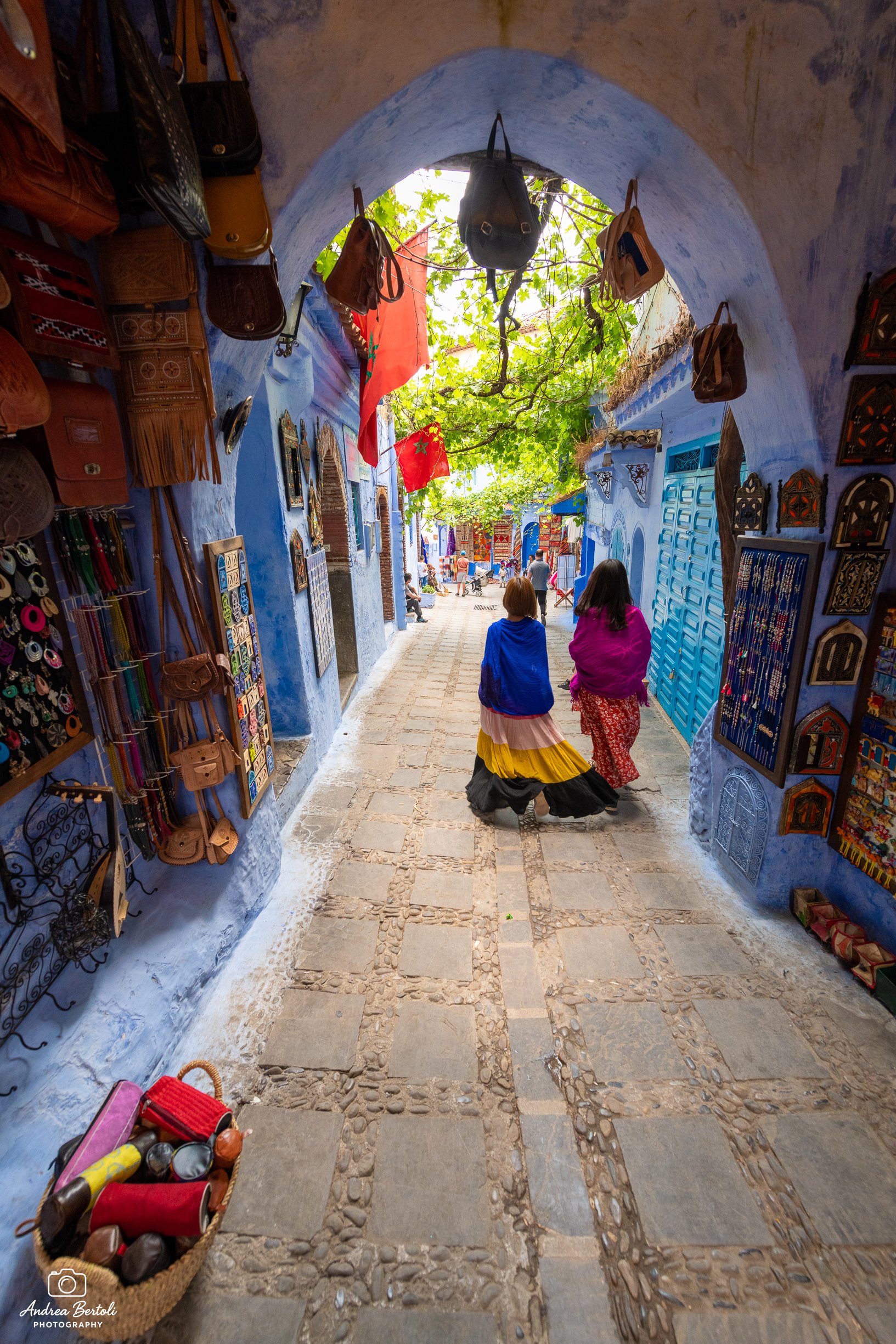 Strada acciottolata all'interno di un vicolo di Chefchaouen e negozi, con persone che passeggiano. Sul lato sinistro, esposizione di borse. Sull'altra parete, decorazioni e oggetti artigianali.