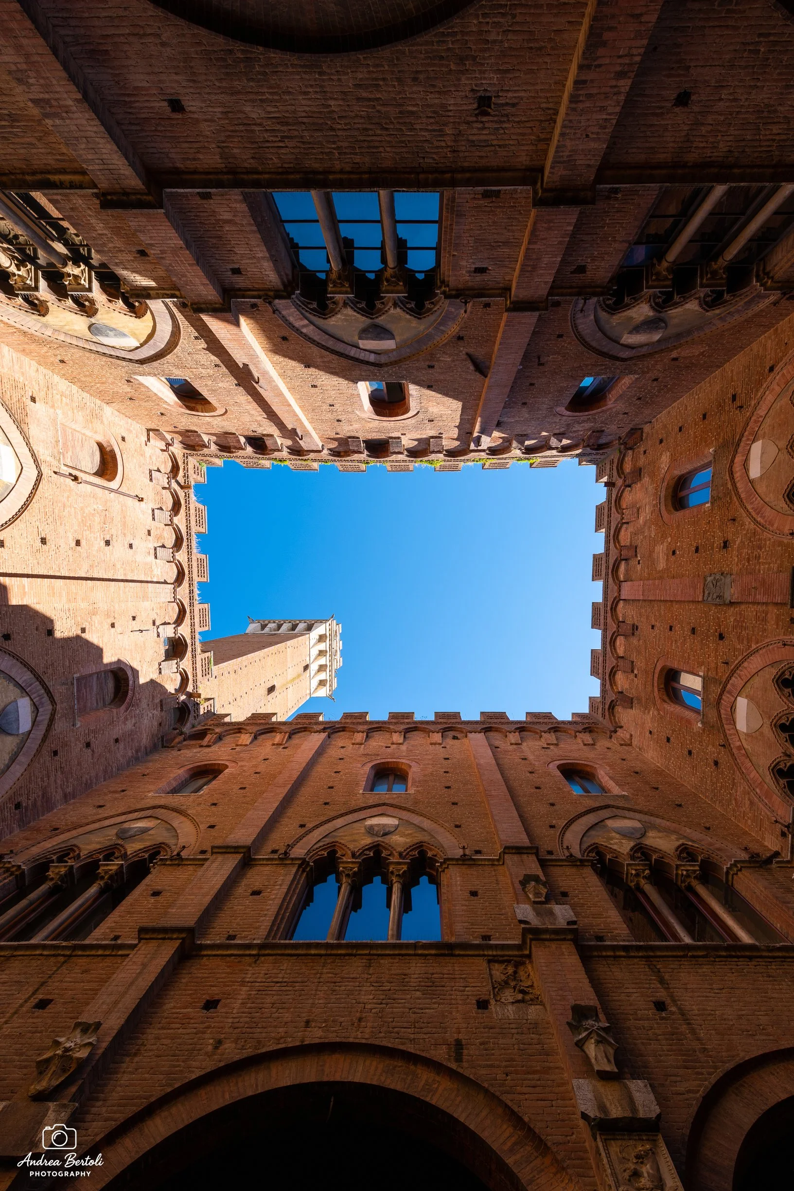 Vista di un cortile interno di un edificio storico senese in mattoni con finestre e archi, visto dal basso verso l'alto con il cielo blu in background.