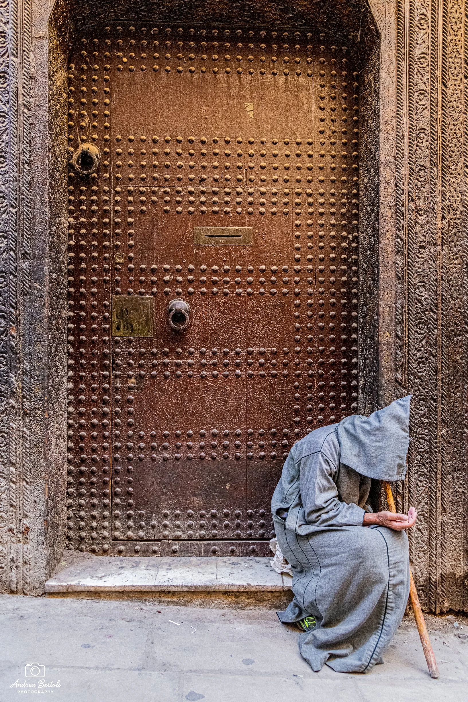 Un mendicante vestito con abiti grigi è inginocchiato davanti a una porta di metallo grande e decorata, mentre chiede la carità, vicino all'entrata di un edificio storico.