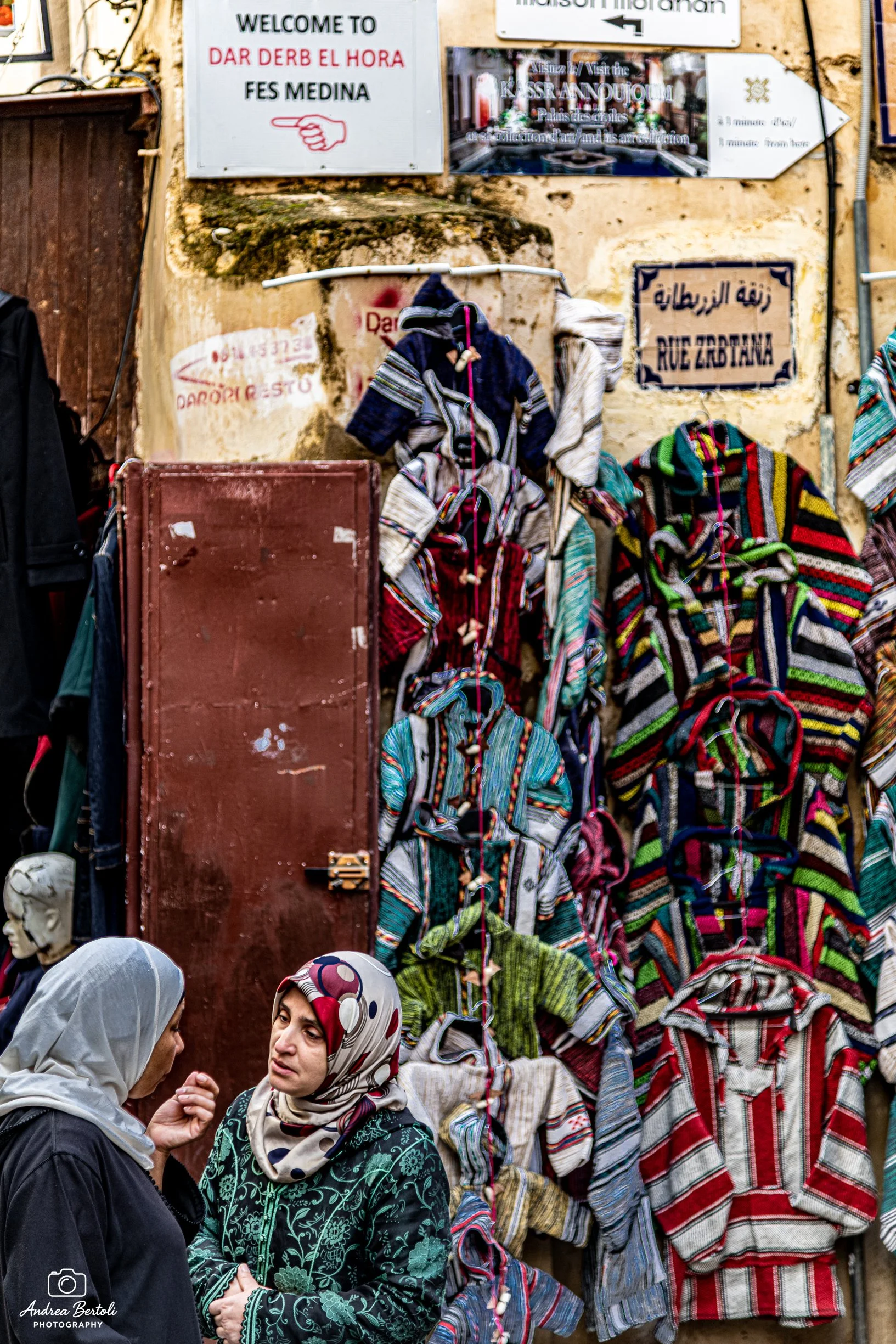 Gruppo di donne arabe che parlano tra loro, alcune con abiti colorati a strisce e altre con hijab, in una piazza affollata. Sullo sfondo, cartelli e muri con scritte in diverse lingue.