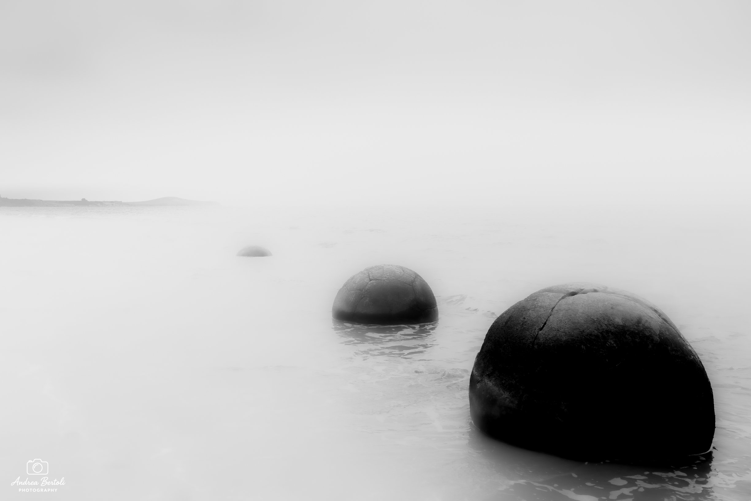 Vista delle Moeraki Boulders in Nuova Zelanda, con grandi pietre rotonde parzialmente sommerse in acqua calma con una nebbia alta e una linea di costa sfocata in lontananza.