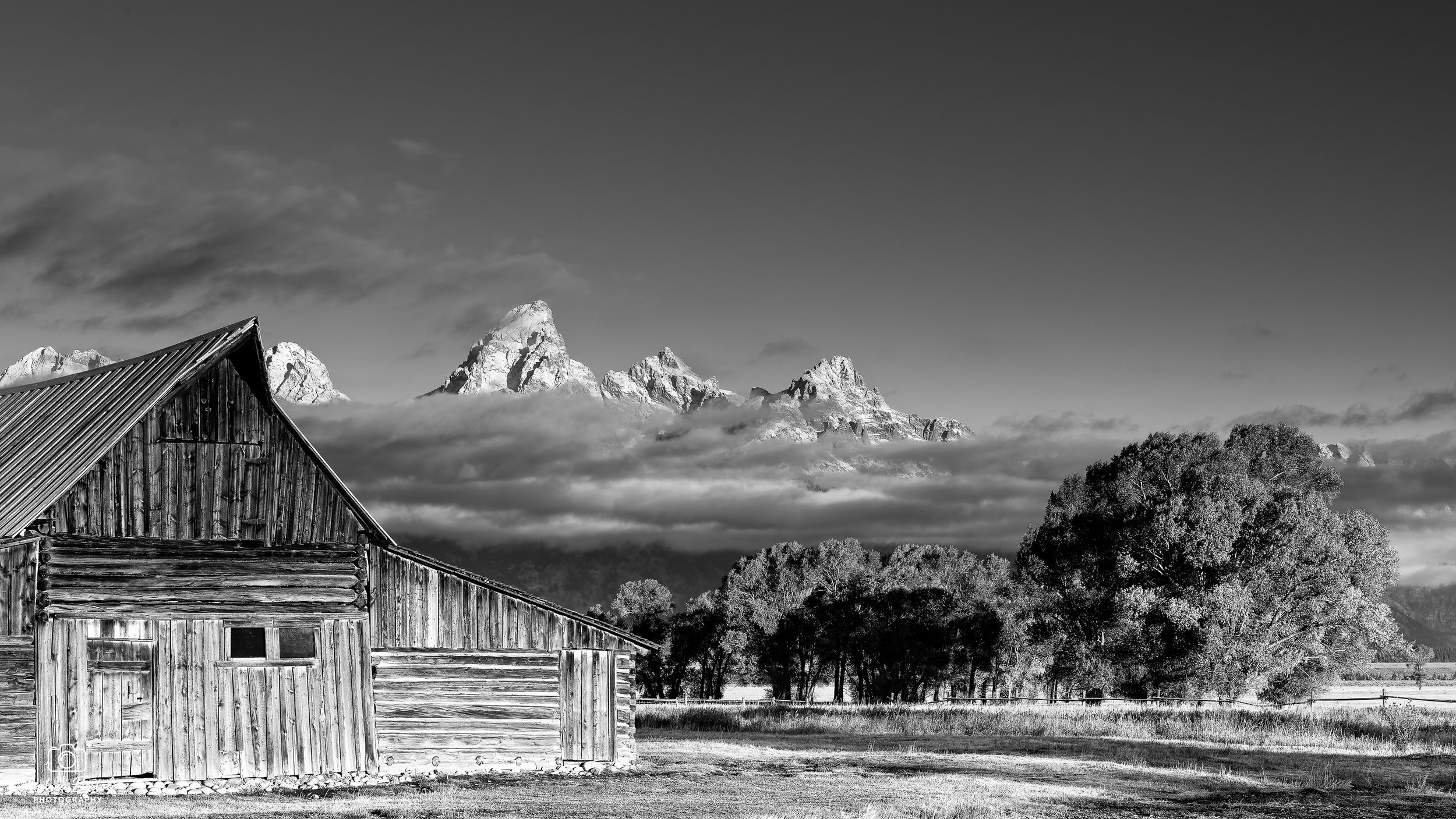 Scena in bianco e nero di un vecchio capannone di legno in primo piano all'interno del Mormon Row Historic District, con montagne e nuvole sullo sfondo.