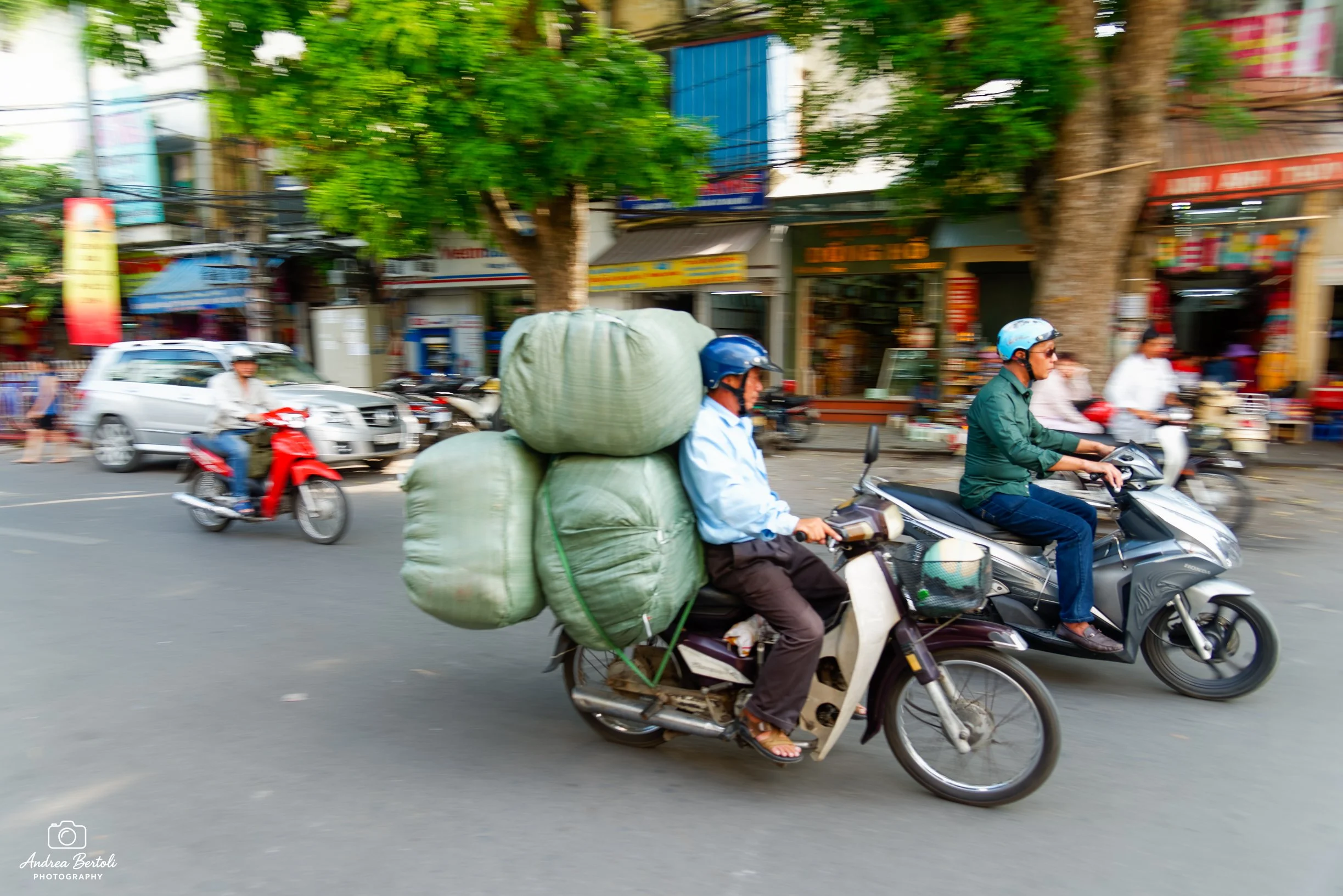 Uomo su scooter con grandi sacchi di tessuto sulle spalle che trasporta oggetti in una strada trafficata con molte persone e veicoli, negozi colorati sullo sfondo.