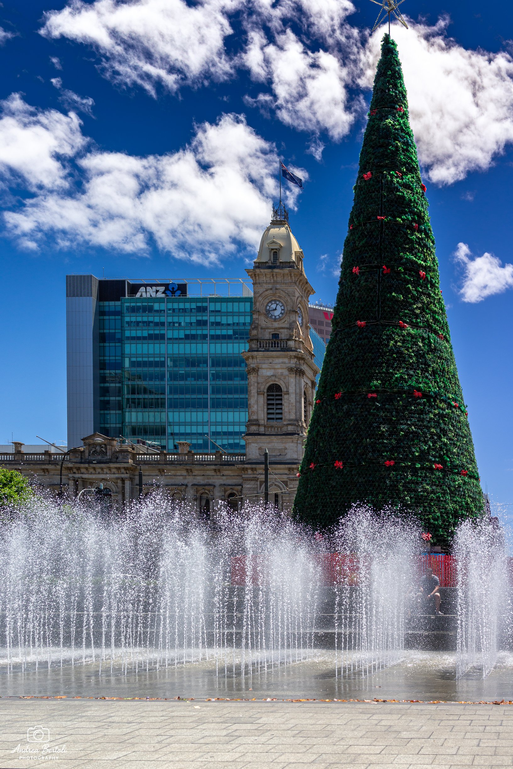 Albero di Natale decorato in una piazza di Adelaide con fontane d'acqua e un edificio con orologio e bandiera sopra.