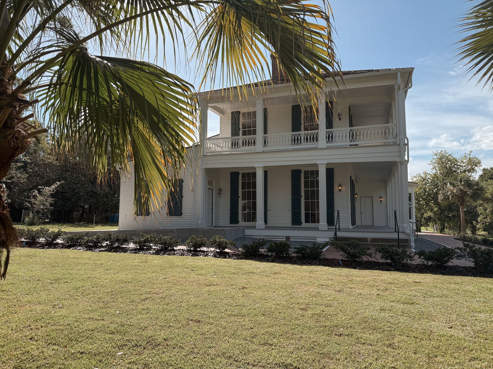 A white, three-story house with dark shutters and a wraparound balcony, situated in a tropical setting with palm trees and a well-maintained lawn.