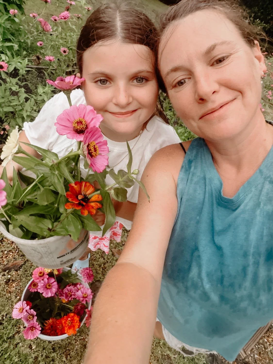 A woman and a young girl taking a selfie outdoors, surrounded by pink and orange flowers in a garden.