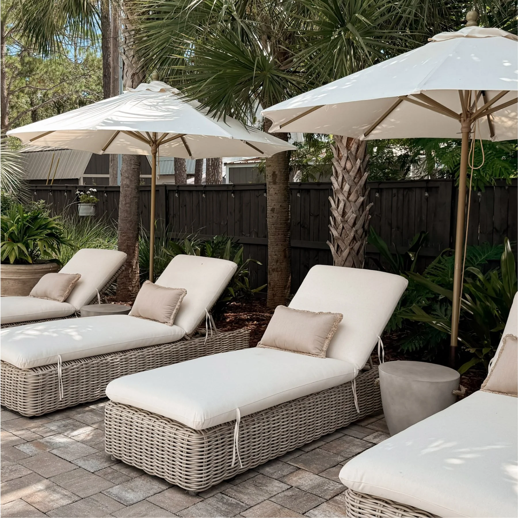 Patio with white wicker lounge chairs with cushions, surrounded by tropical plants, trees, and large white umbrellas, on a stone-paved surface.