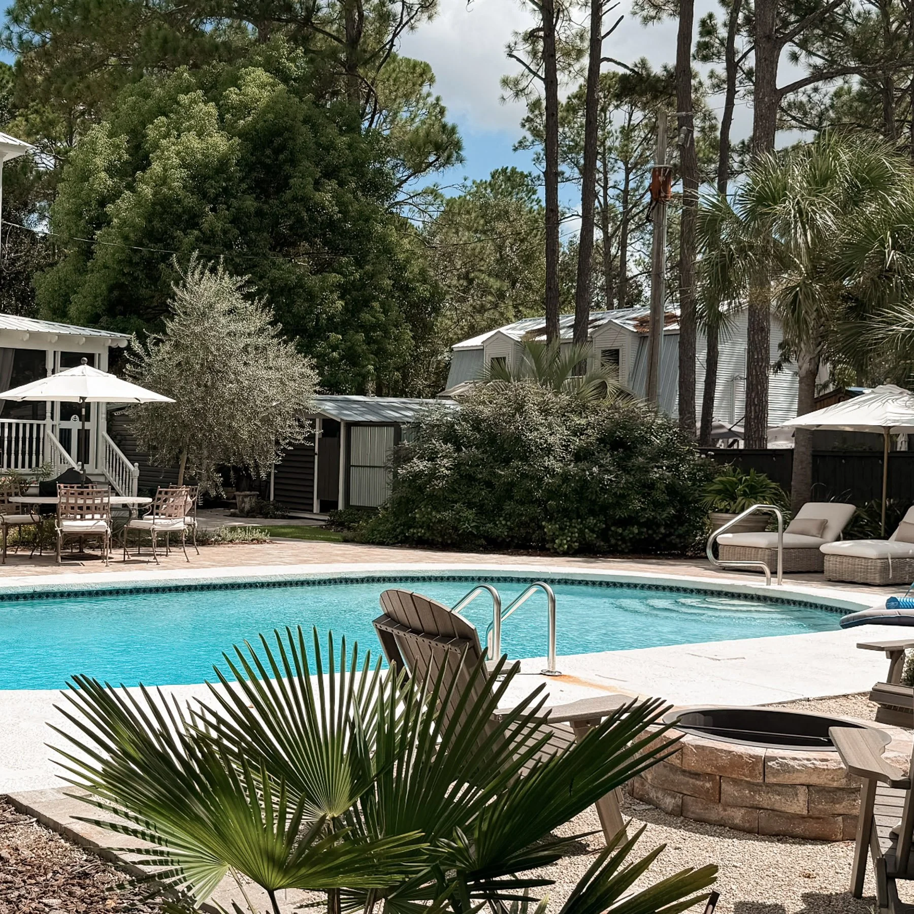 A backyard swimming pool area with patio furniture, umbrellas, and landscaping including palm trees and other greenery, with houses and tall trees in the background.