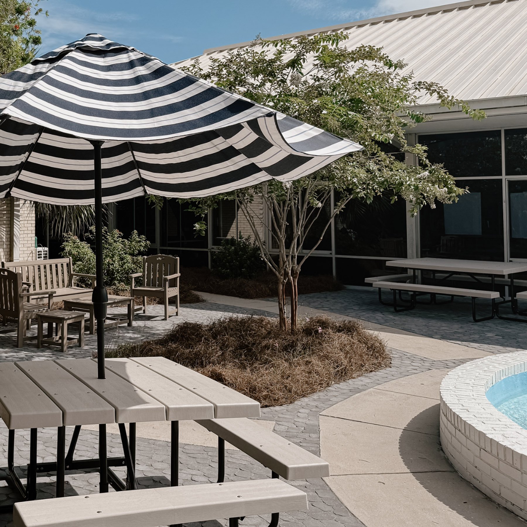 Outdoor patio area with white picnic tables, a striped black and white umbrella, small trees, and a screened-in porch in the background on a sunny day.
